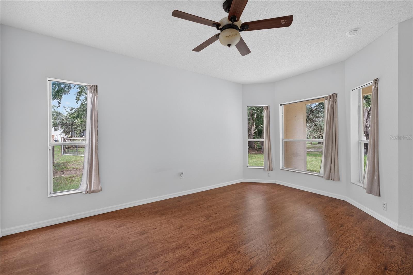 Vacant living room with wood floors, a ceiling fan, and several windows.