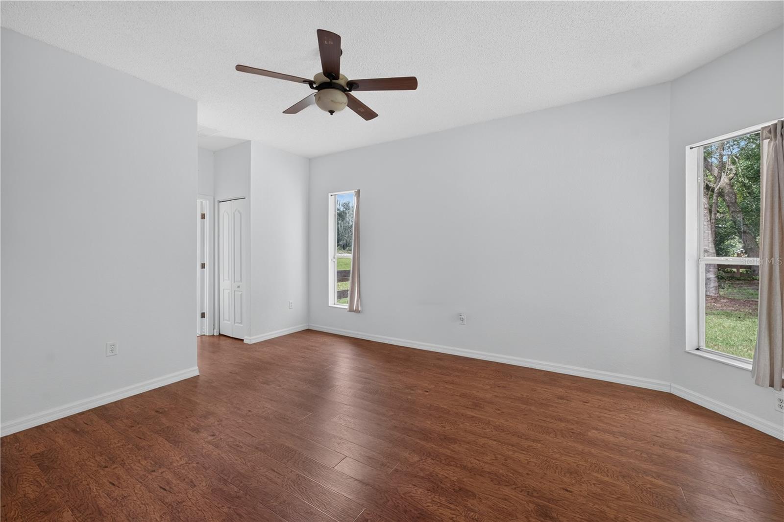 Empty room with reddish-brown flooring, a ceiling fan, and windows overlooking trees.