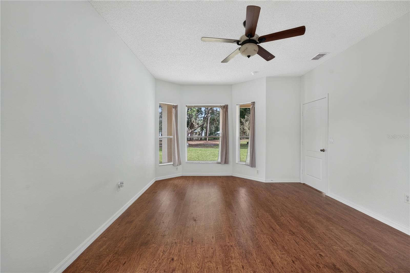 Empty room with hardwood floors, three windows overlooking a lawn, and a ceiling fan.