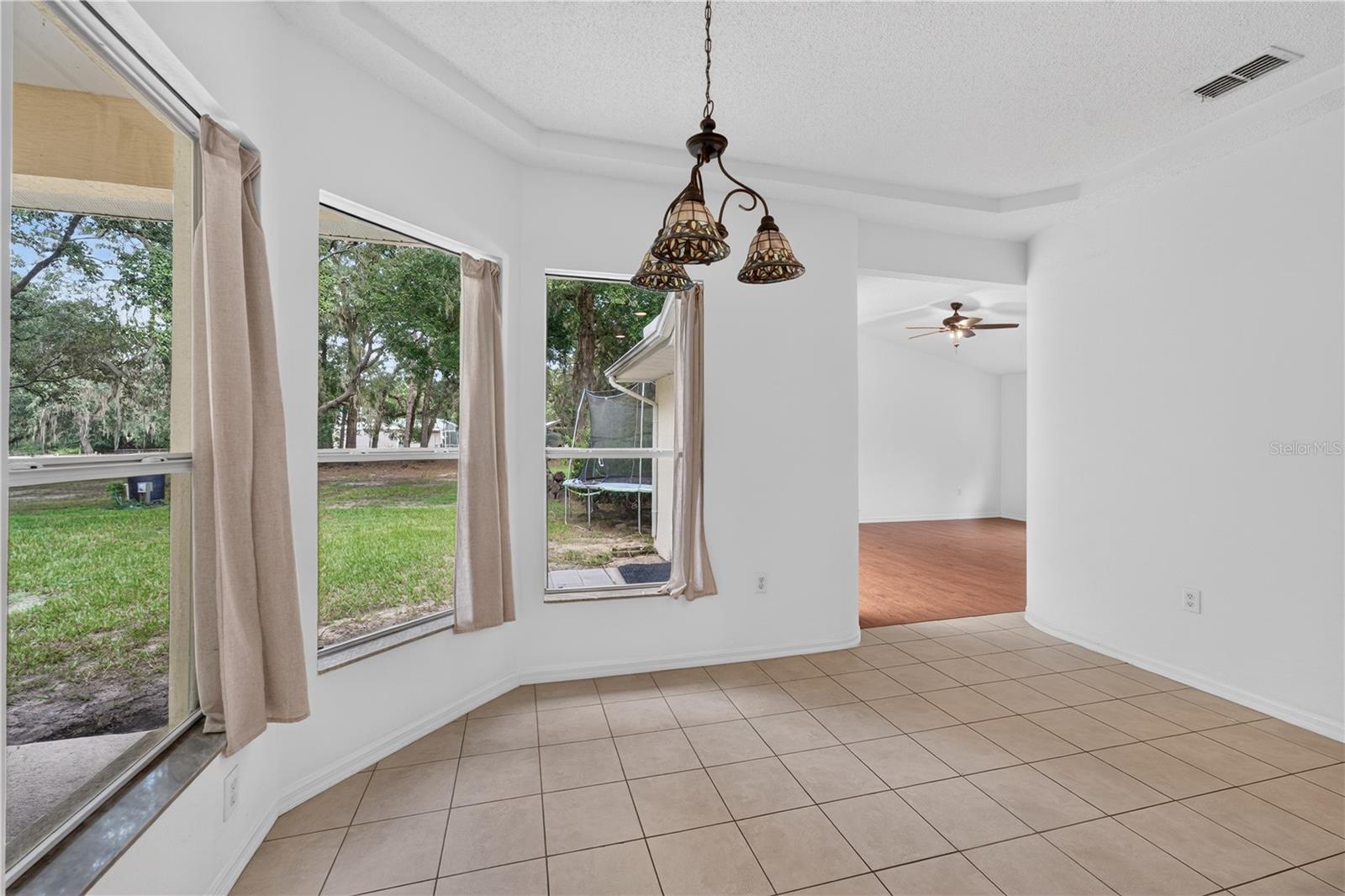 Dining area with tiled floors, three curtained windows overlooking the yard, and a decorative chandelier.