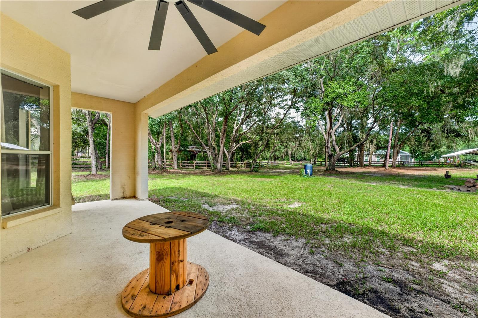 Covered patio with a ceiling fan and spool table overlooking a tree-lined grassy backyard.