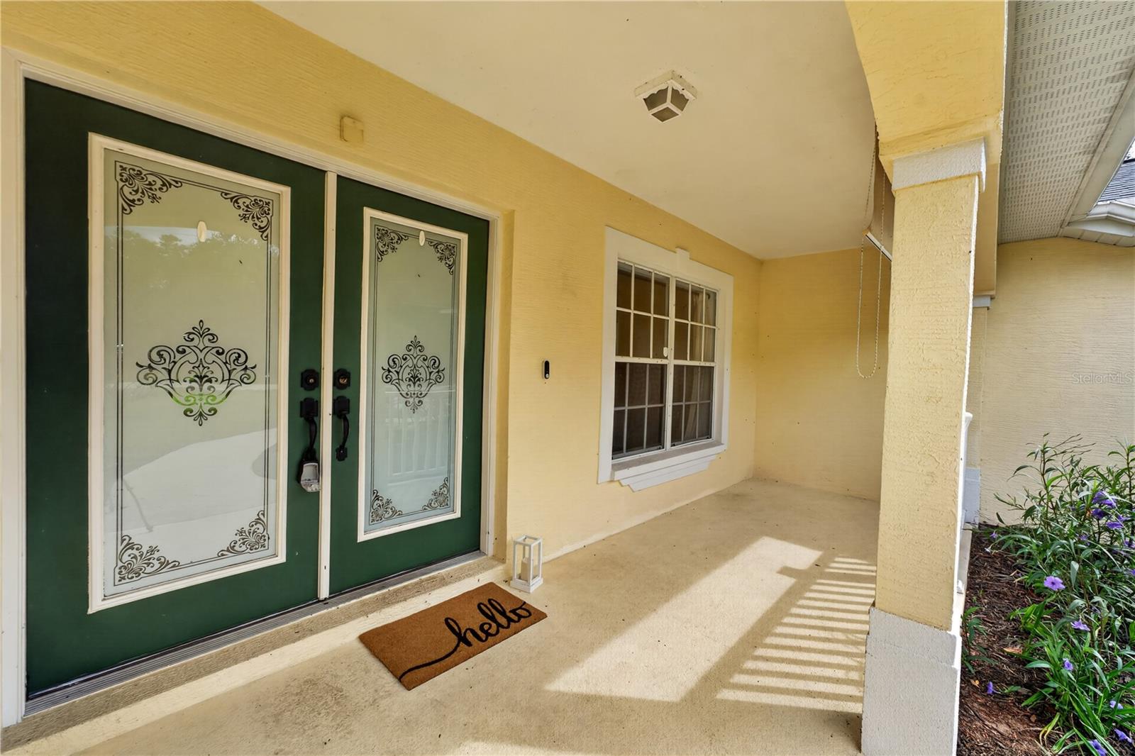 Covered front porch with decorative green double doors, a white-trimmed window, and a hello doormat.