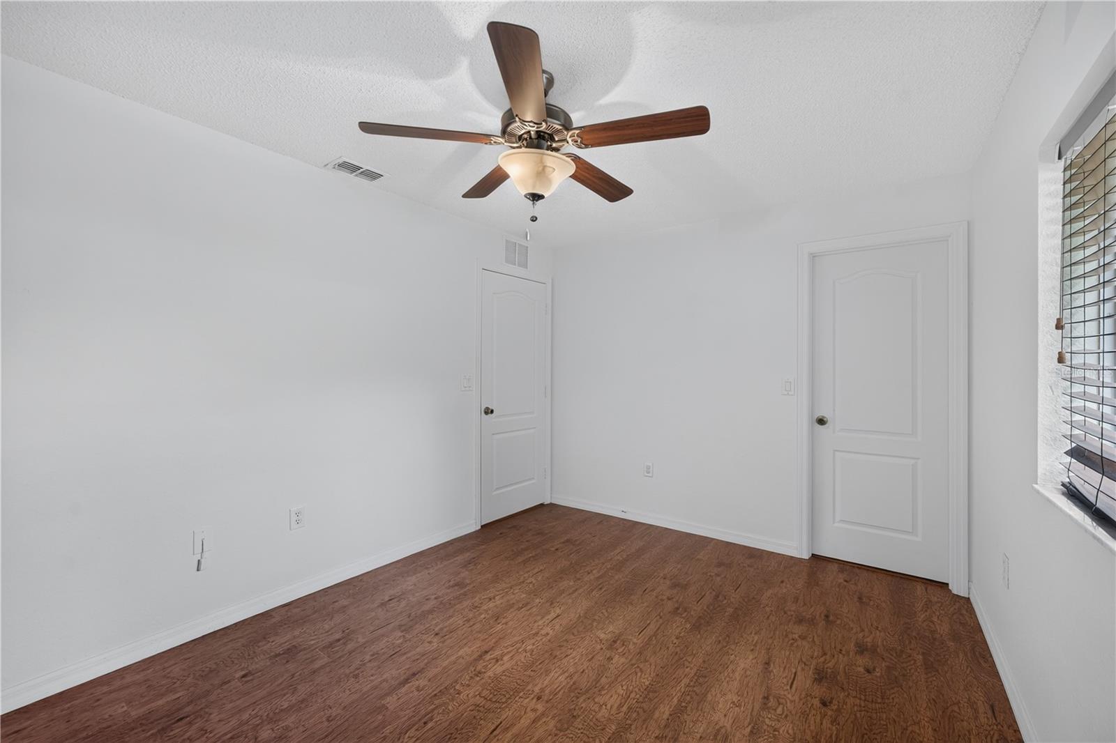Vacant room with reddish wood flooring, a ceiling fan, two doors, and a window with blinds.