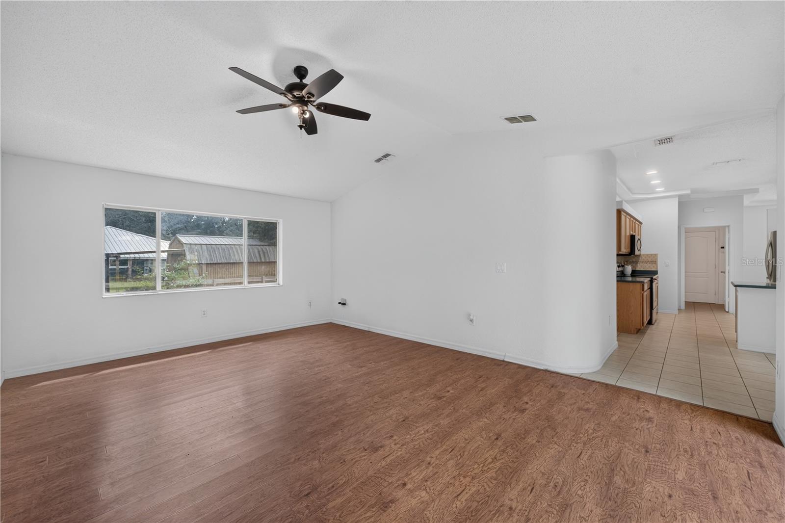 Vacant living room with wood-look flooring, a wide window, and the kitchen visible to the right.