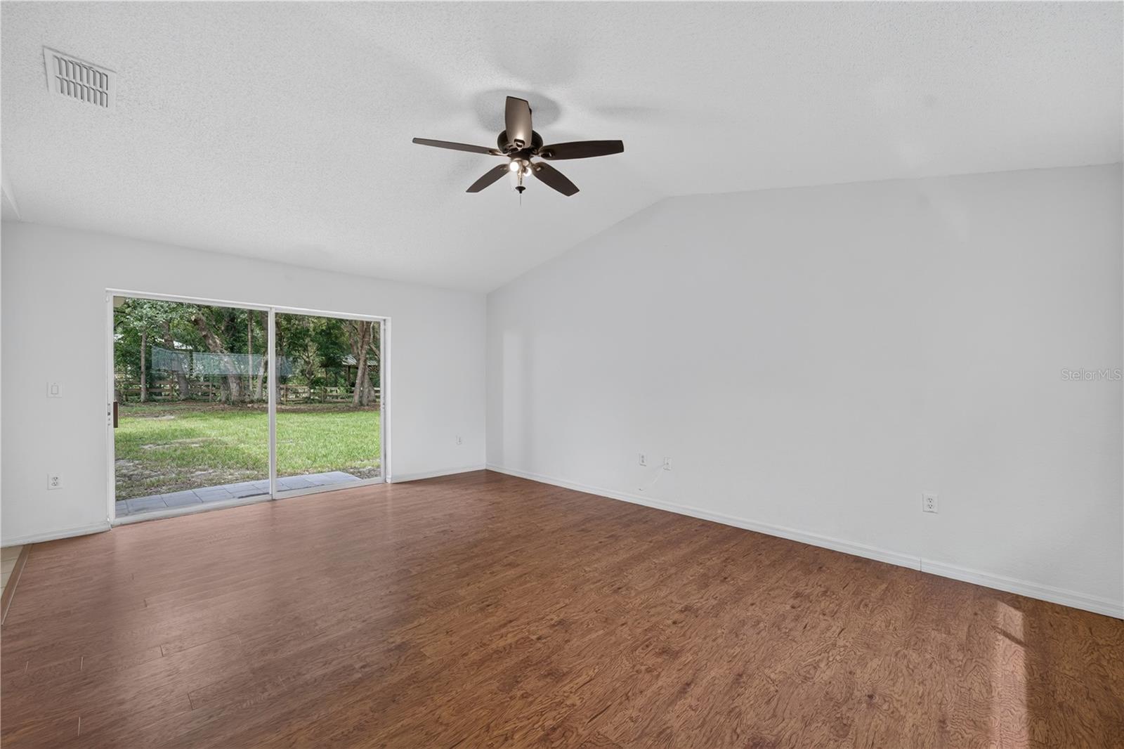Empty living room with wood-look floors, ceiling fan, and sliding glass doors facing a grassy backyard.