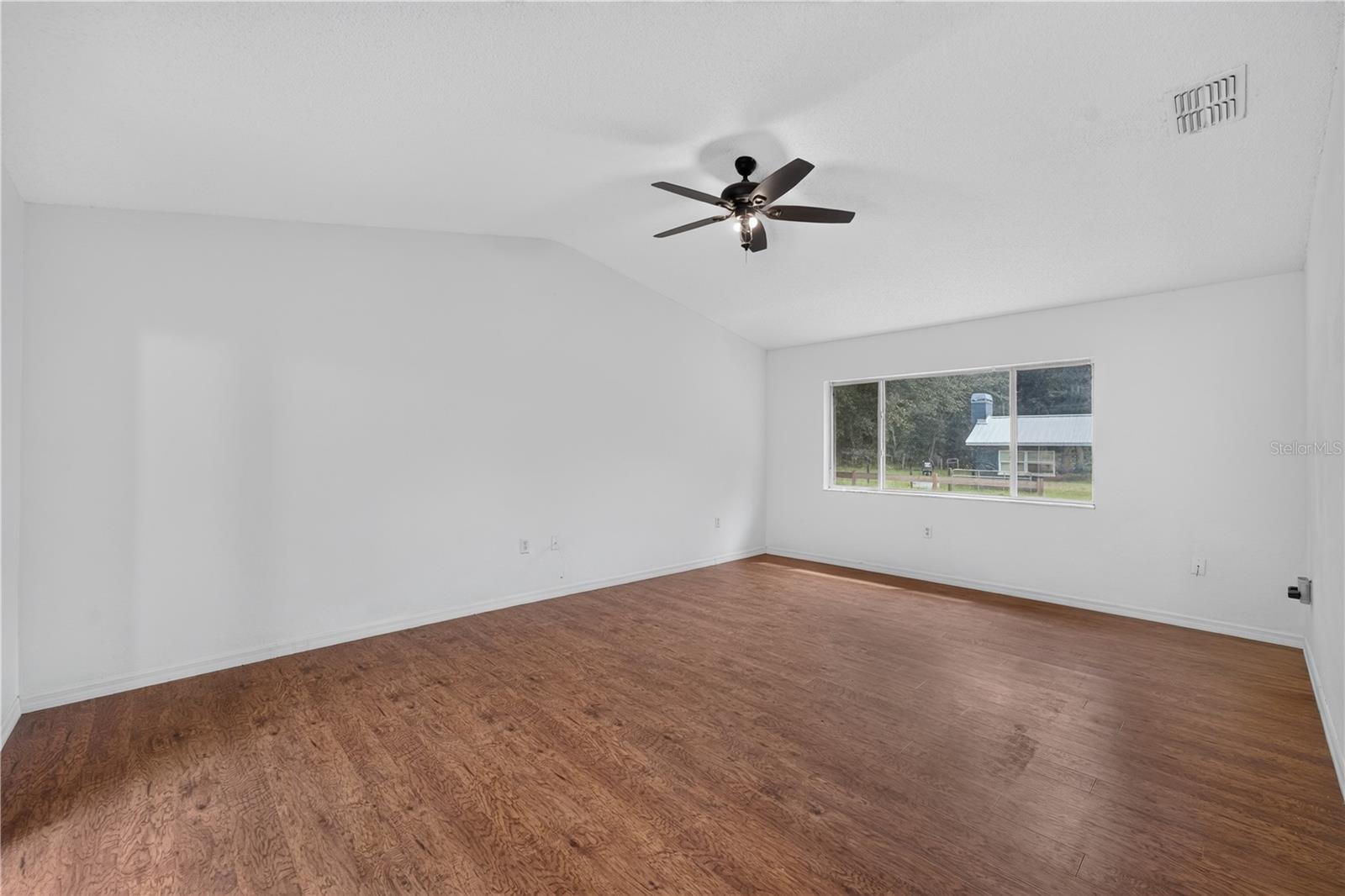 Empty room with brown wood-look flooring, a ceiling fan, and a wide window overlooking trees.