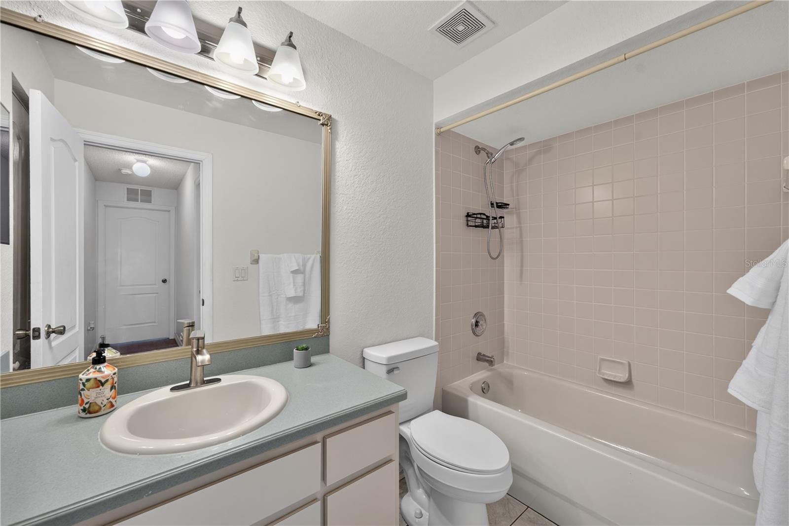 Bathroom with a blue-green vanity counter, large mirror, toilet, and tub with tiled shower surround.