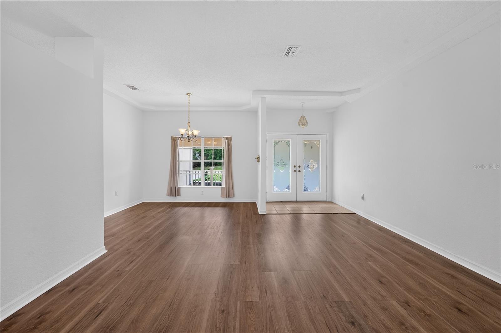 Empty living and dining area with brown flooring, a curtained window, and double glass doors.