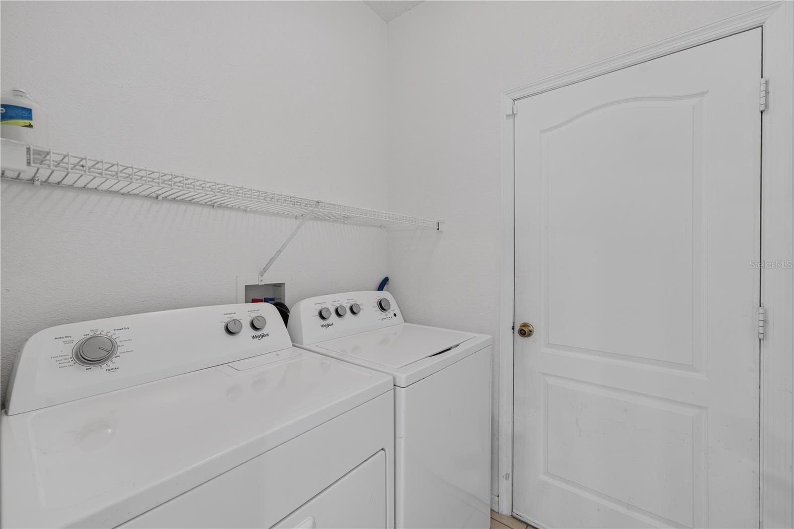 Laundry room with white washer and dryer beneath a wire shelf beside a paneled door.
