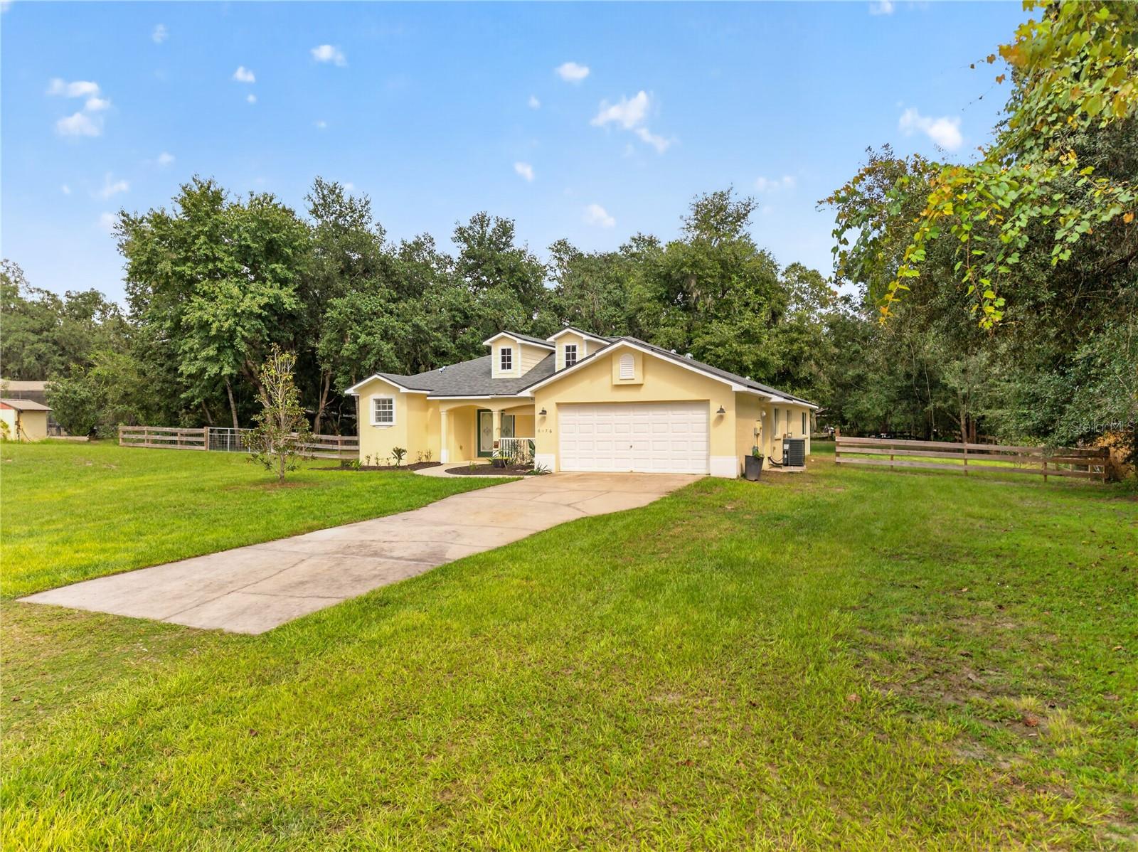 Yellow house with a two-car garage, long driveway, broad lawn, and wooden fencing.