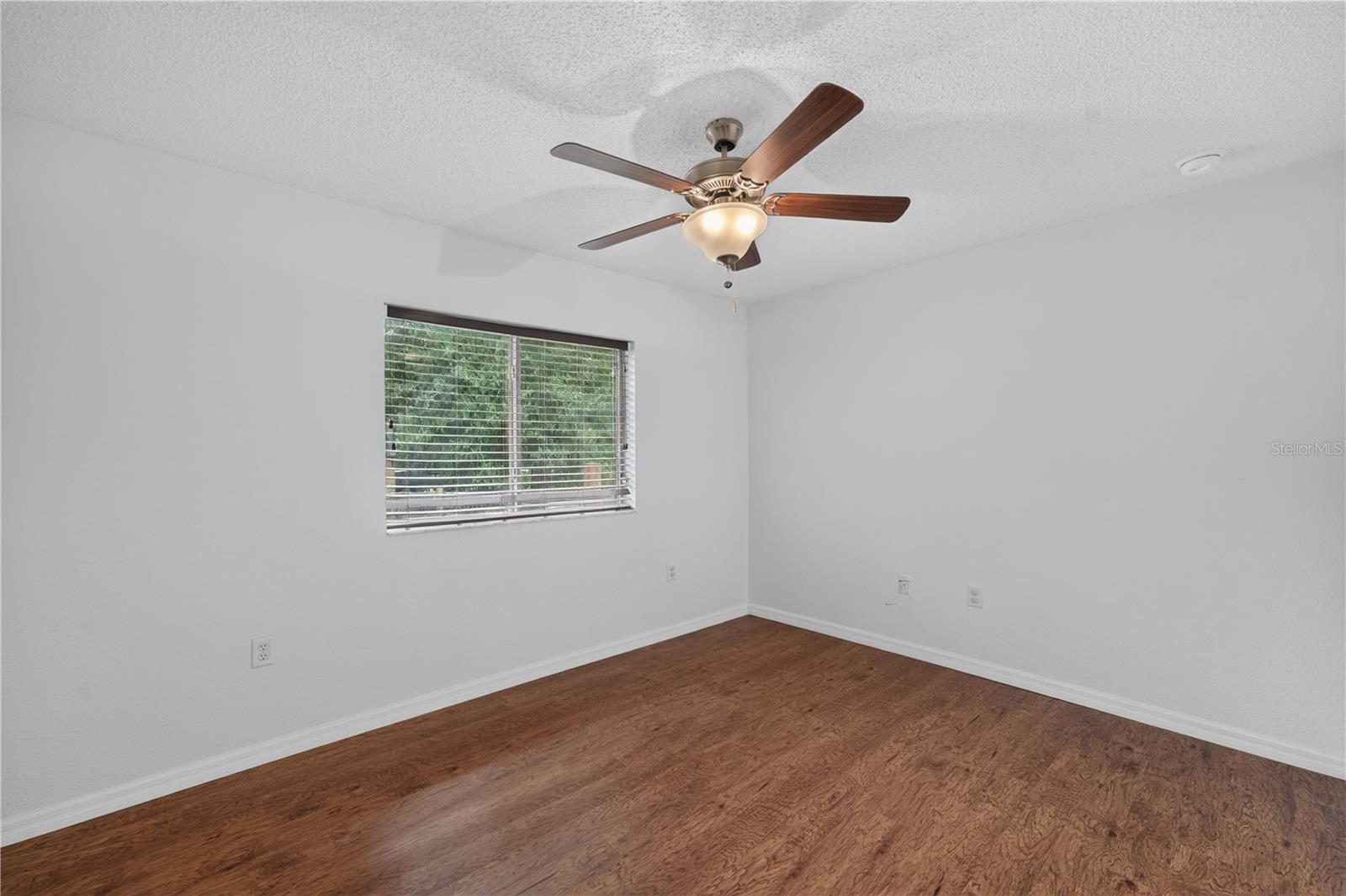 Empty bedroom with wood flooring, window blinds, and a ceiling fan with light.