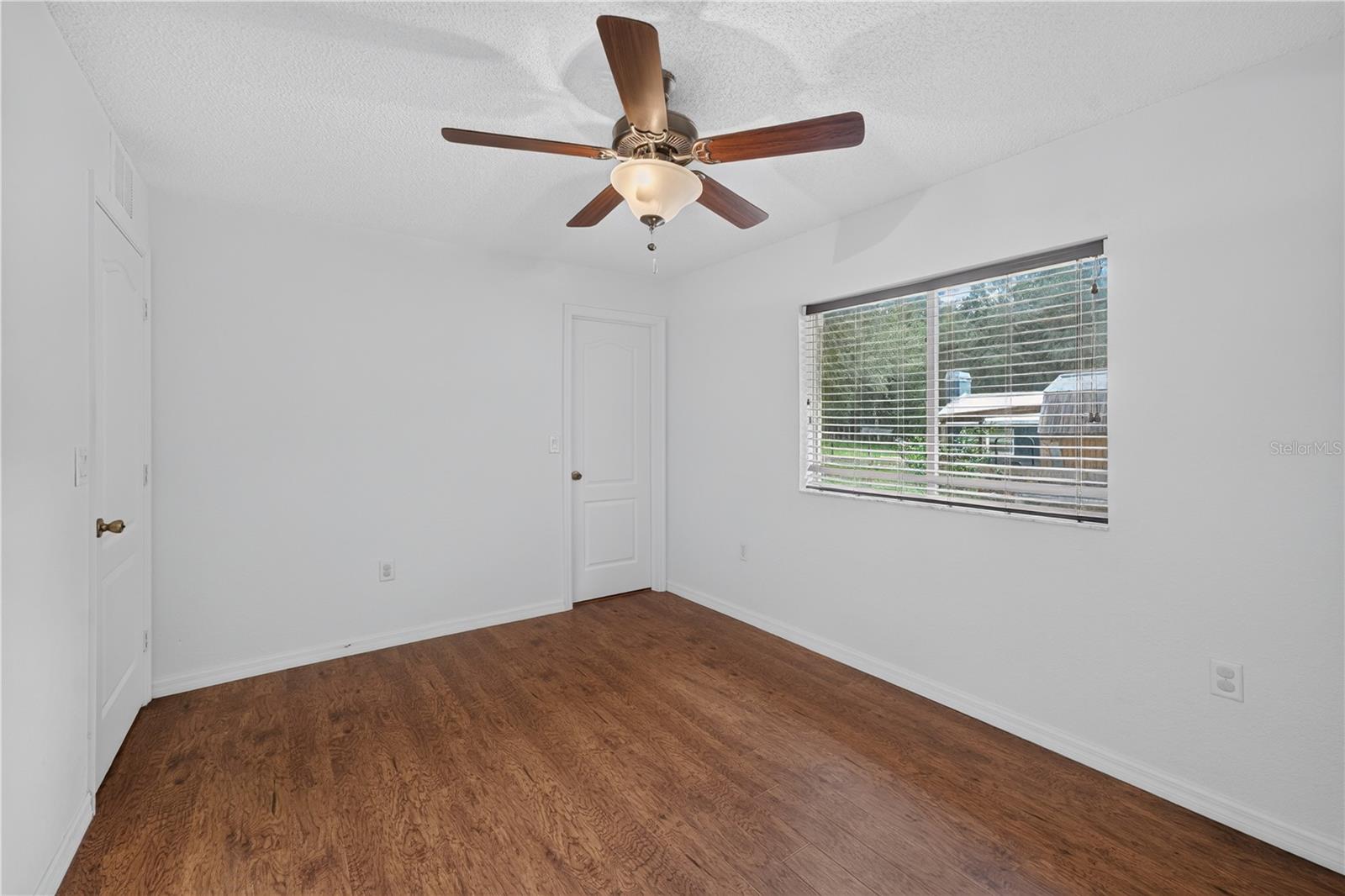 Empty room with brown wood-look flooring, a ceiling fan, and a window with blinds.