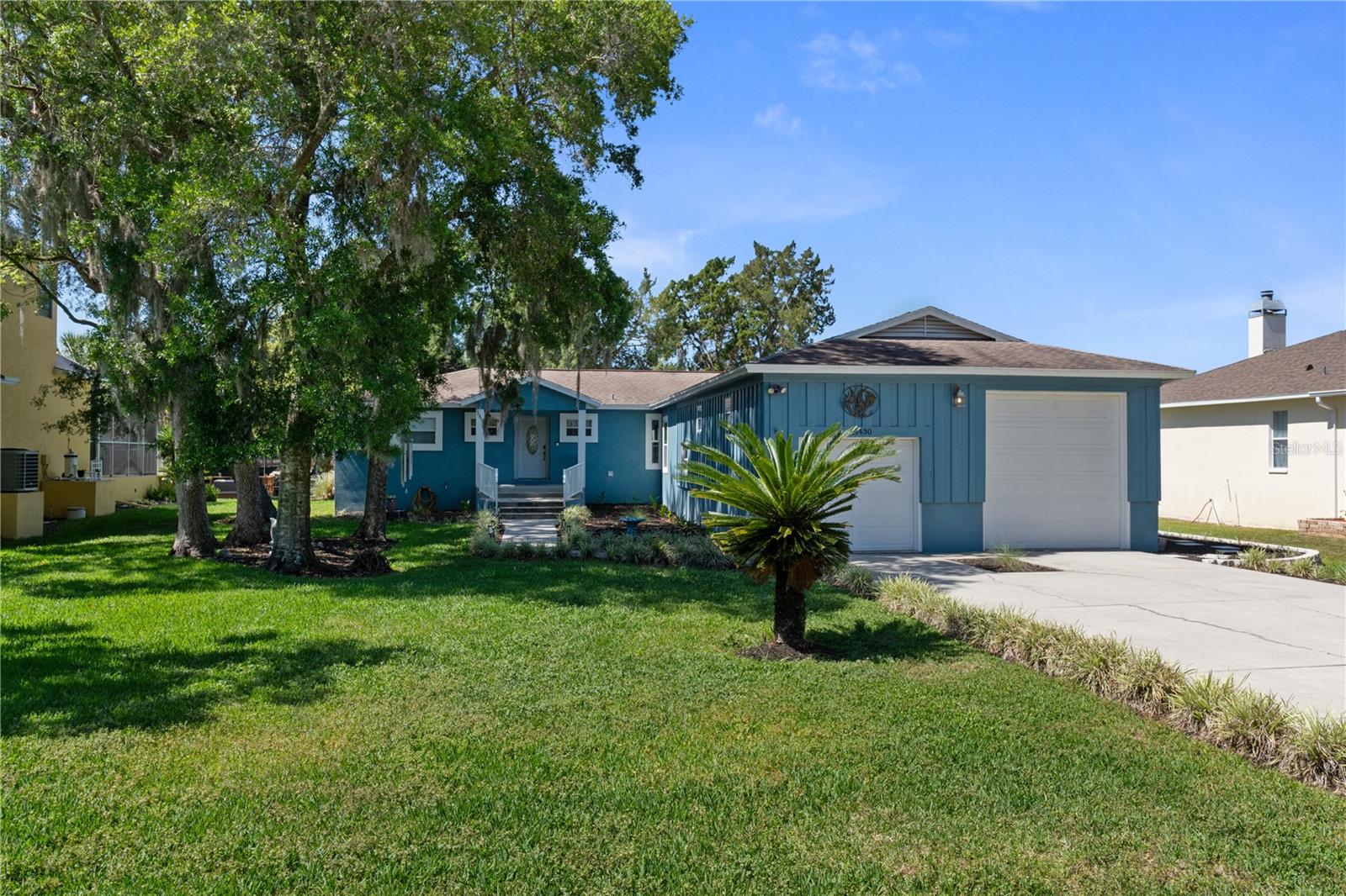 Blue-sided home with a two-car garage, broad driveway, front lawn, and mature trees.