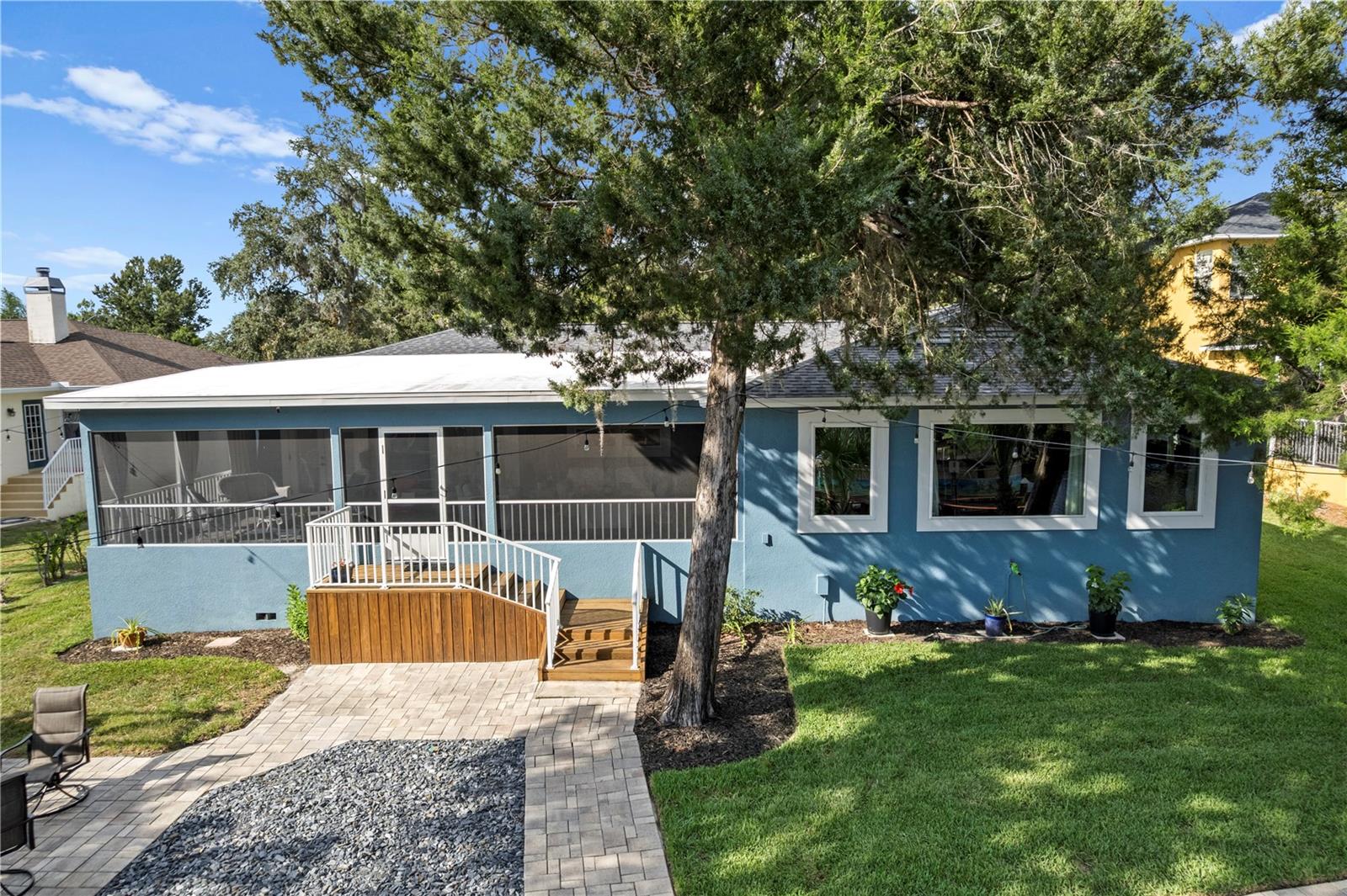 Blue house with a screened porch and white-railed deck beside a paved patio and large tree.