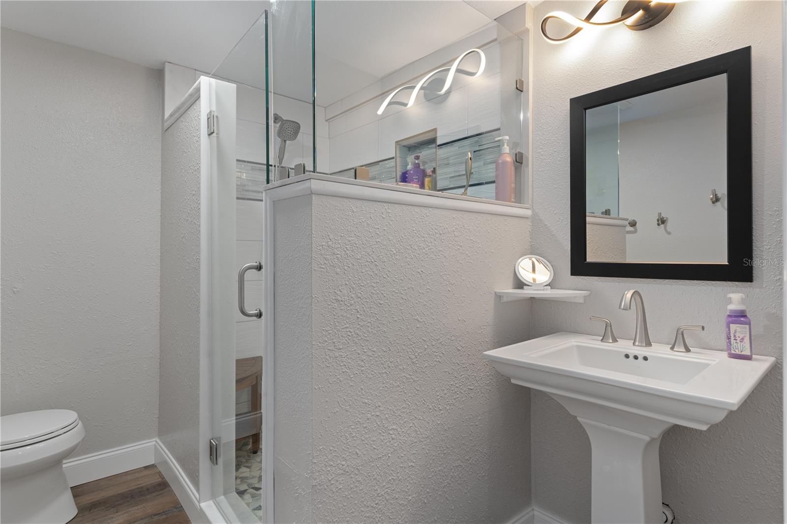 Bathroom with glass-enclosed shower, pedestal sink, and black-framed mirror.