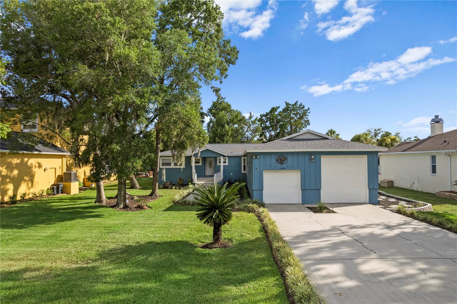 Blue single-story home with white garage doors, a broad driveway, and mature trees.