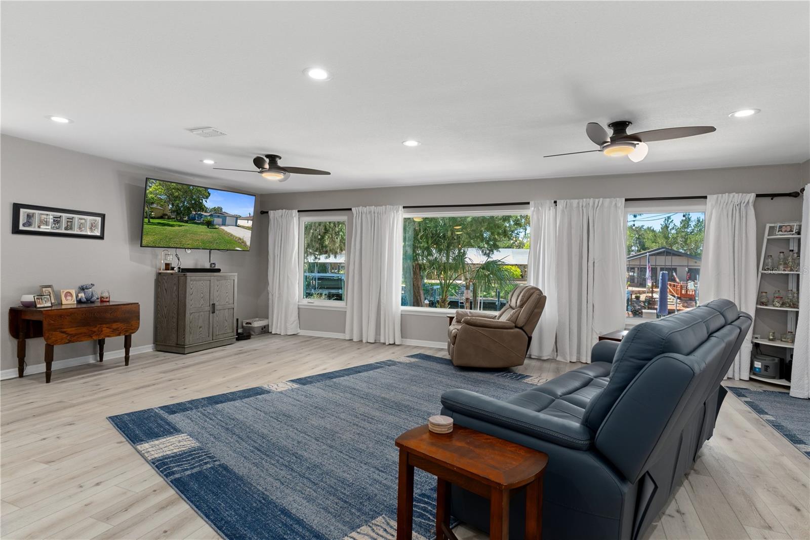 Furnished living room with a blue reclining sofa, broad curtained windows, and pale wood flooring.