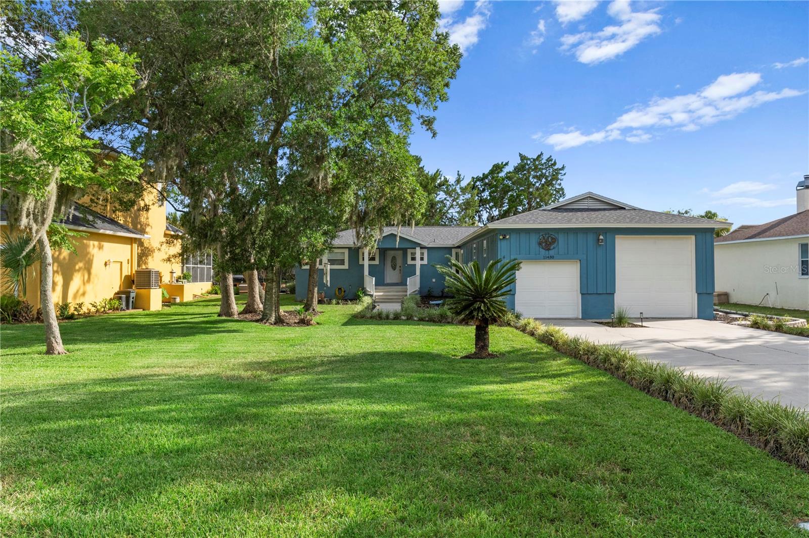 Blue single-story house with a two-car garage, broad lawn, and large shade trees.