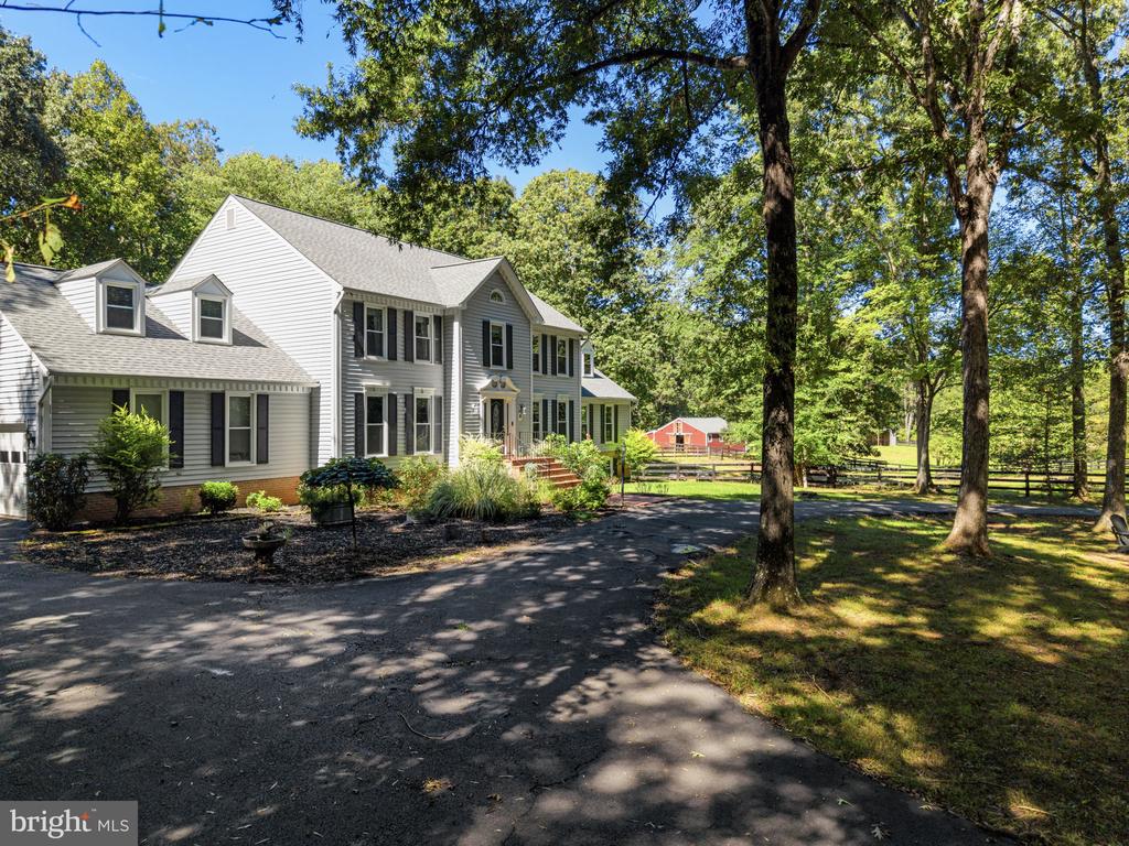 Two-story white home beside a wooded driveway, with fenced pasture and a red barn in the distance.