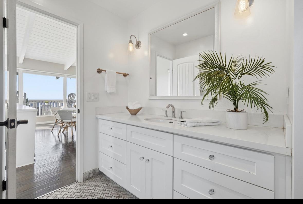 Bright bathroom vanity with brass fixtures and a potted palm, opening onto a balcony with an ocean glimpse.