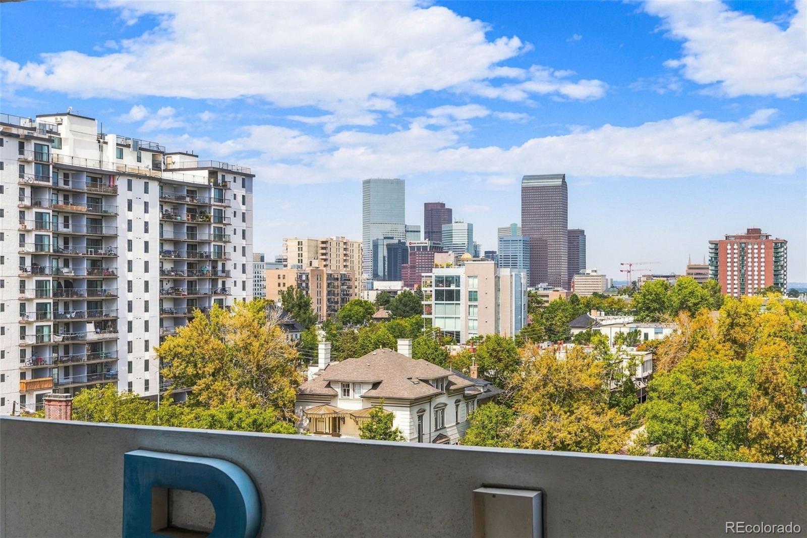 Downtown Denver skyline beyond leafy trees and residential buildings, viewed from a balcony.