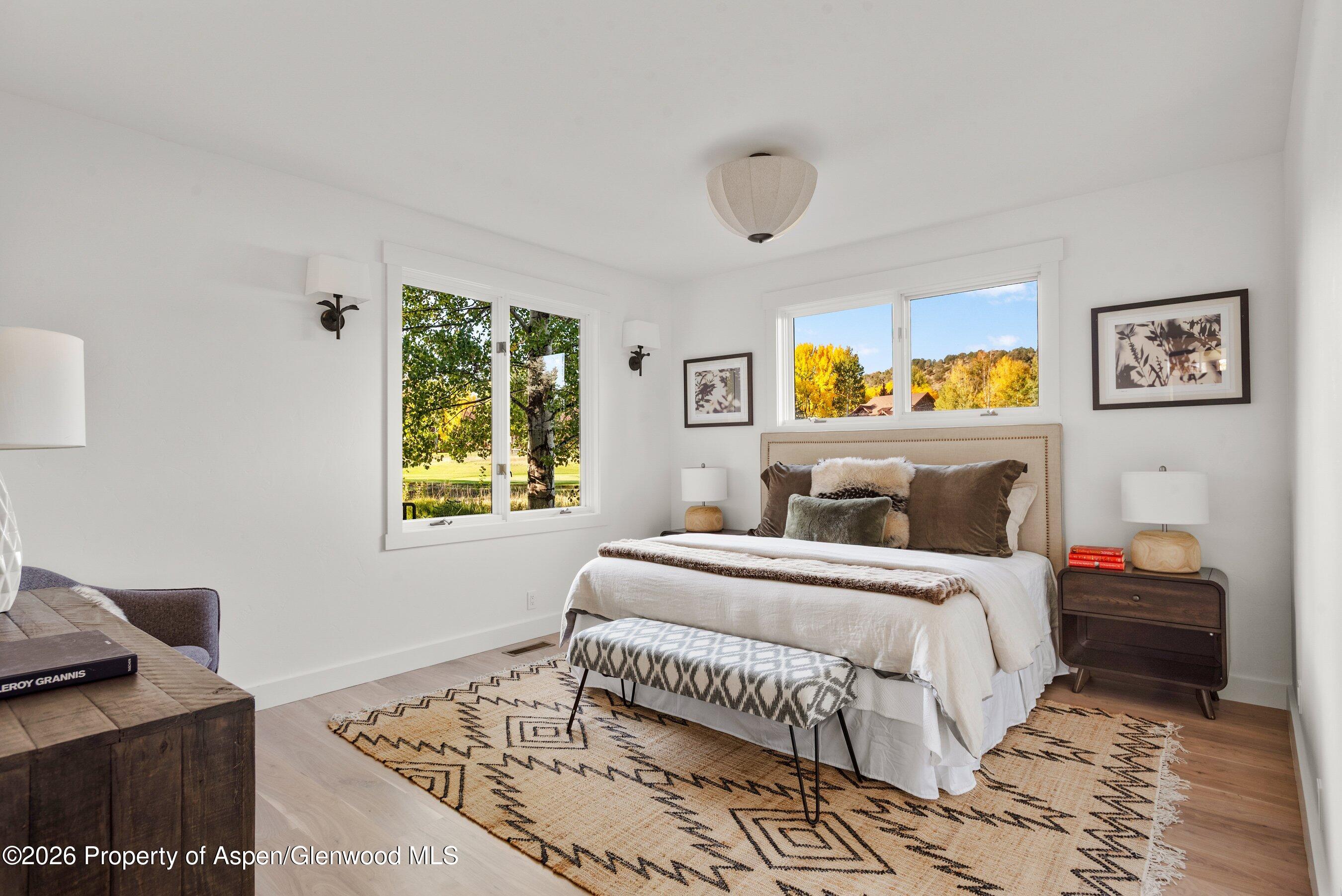 Bedroom with a white bed, patterned rug, and windows overlooking autumn trees.