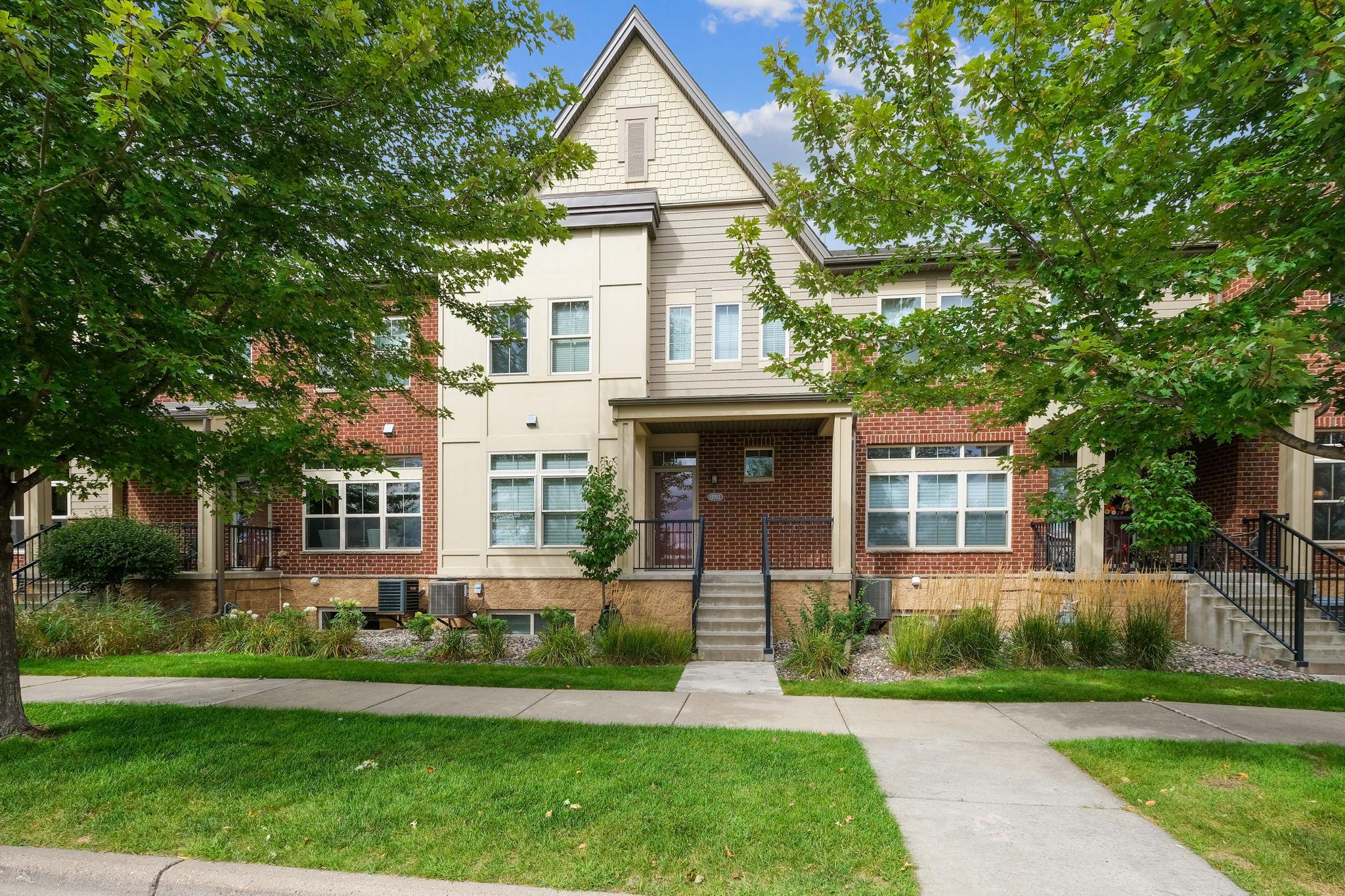 Townhouse exterior with a brick facade, central entry steps, landscaped lawn, and mature trees.