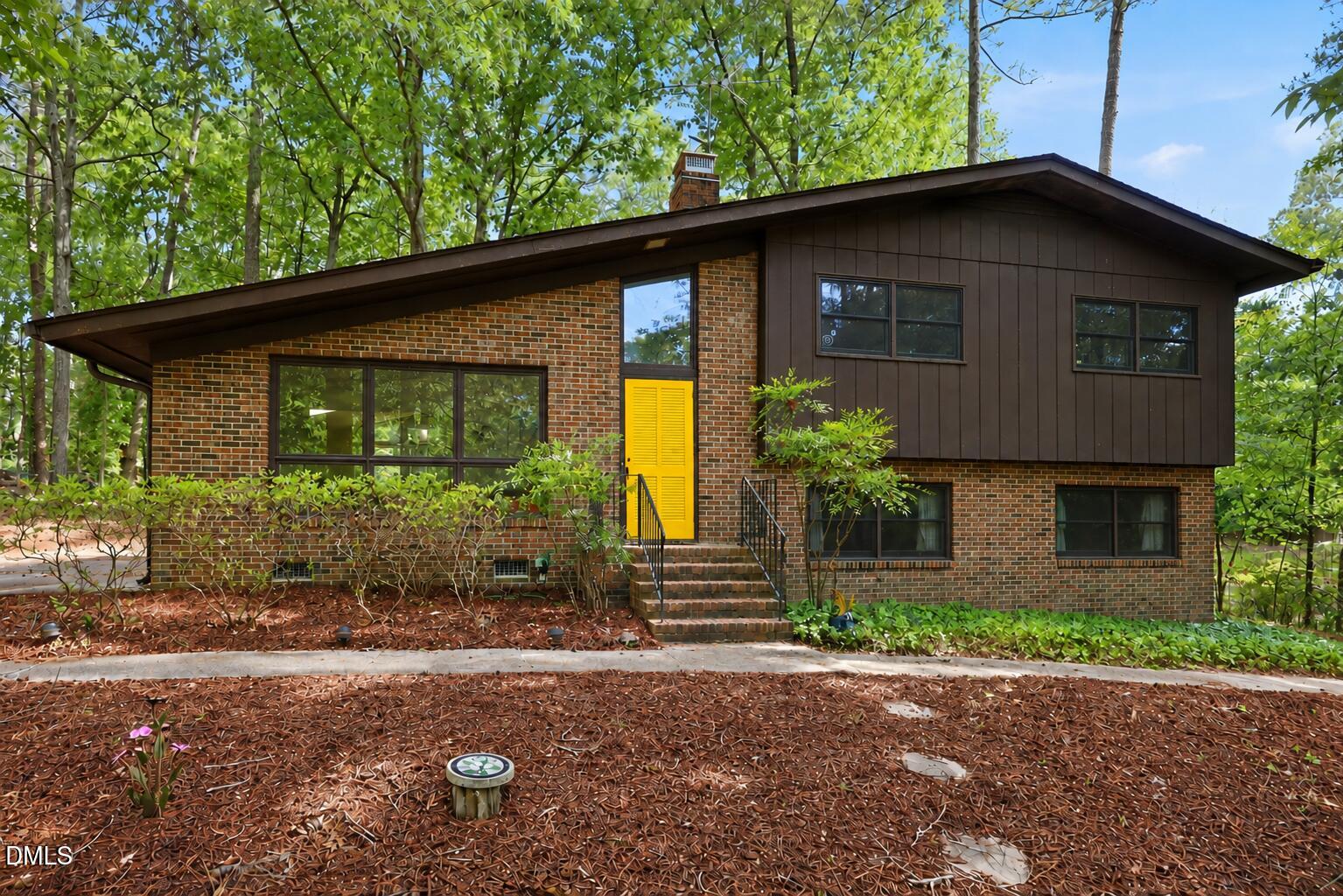 Brick-and-brown-sided house with a bright yellow front door, brick steps, and mature trees.