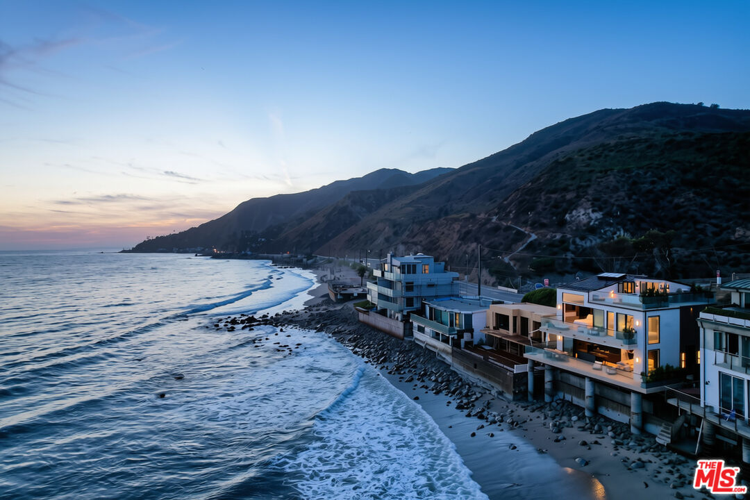 Oceanfront Malibu homes at dusk, with waves, rocky shoreline, and mountains.