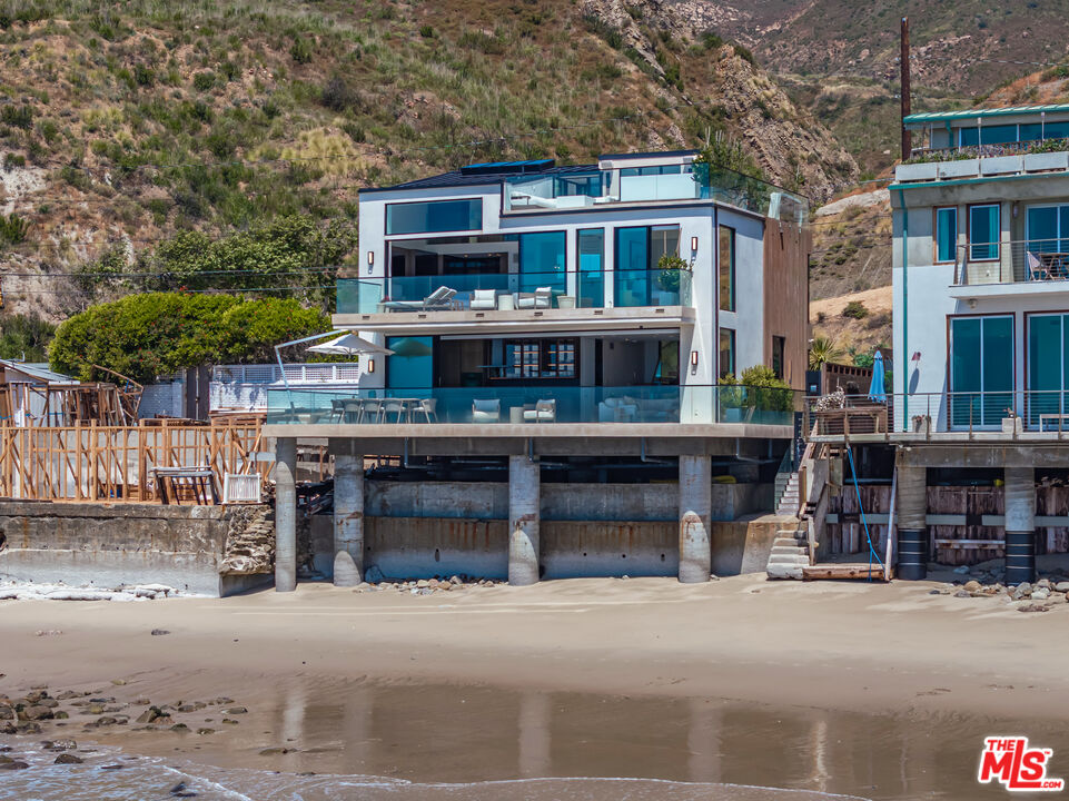 Modern glass-fronted beachfront home raised on concrete pilings, with furnished terraces and a hillside behind it.