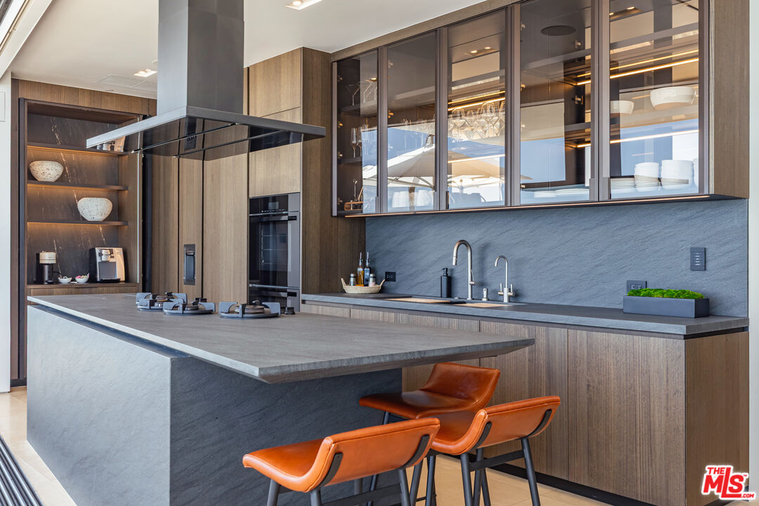 Modern kitchen with a large stone island, orange leather stools, and glass-front wood cabinets.