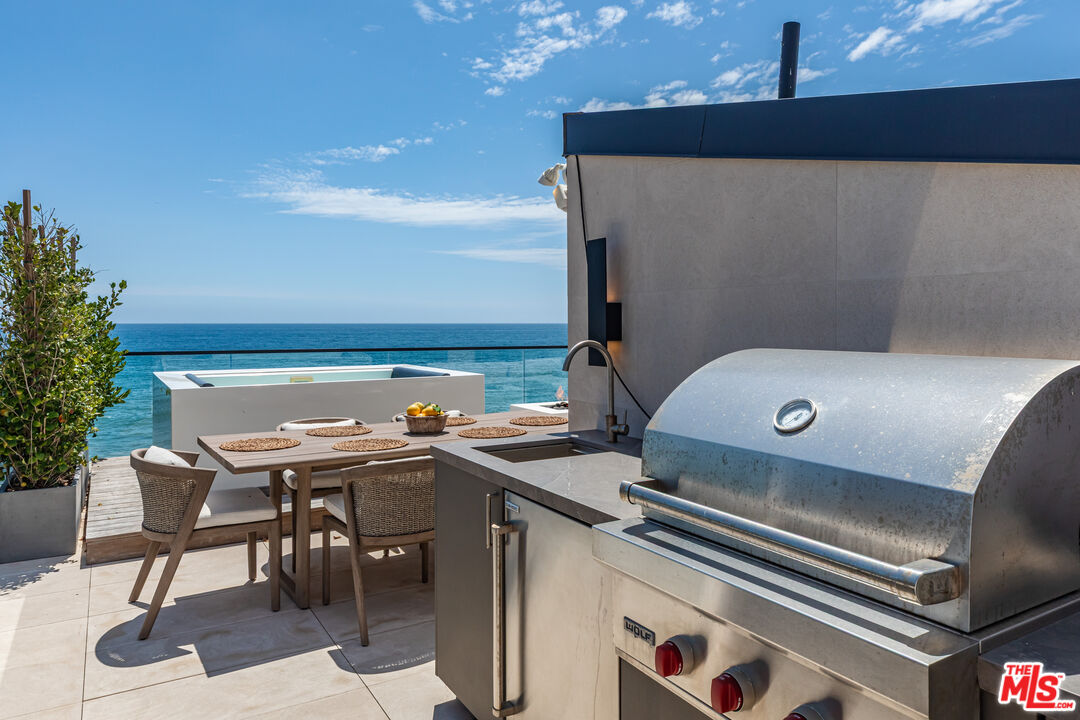 Oceanfront terrace with stainless-steel grill, dining table, and hot tub overlooking the water.