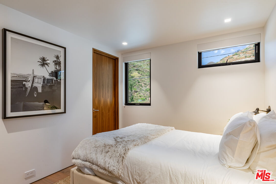 Bedroom with a white bed, framed beach photograph, and windows overlooking a hillside.