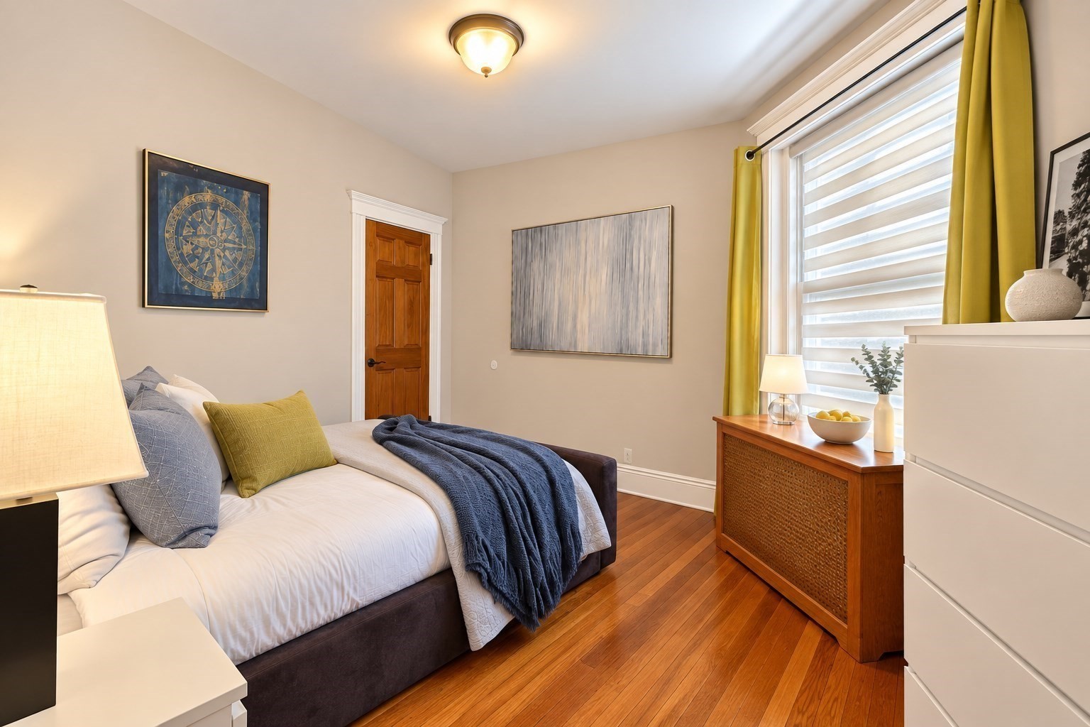 Staged bedroom with a white bed, navy throw, hardwood floors, and a large window with yellow curtains.