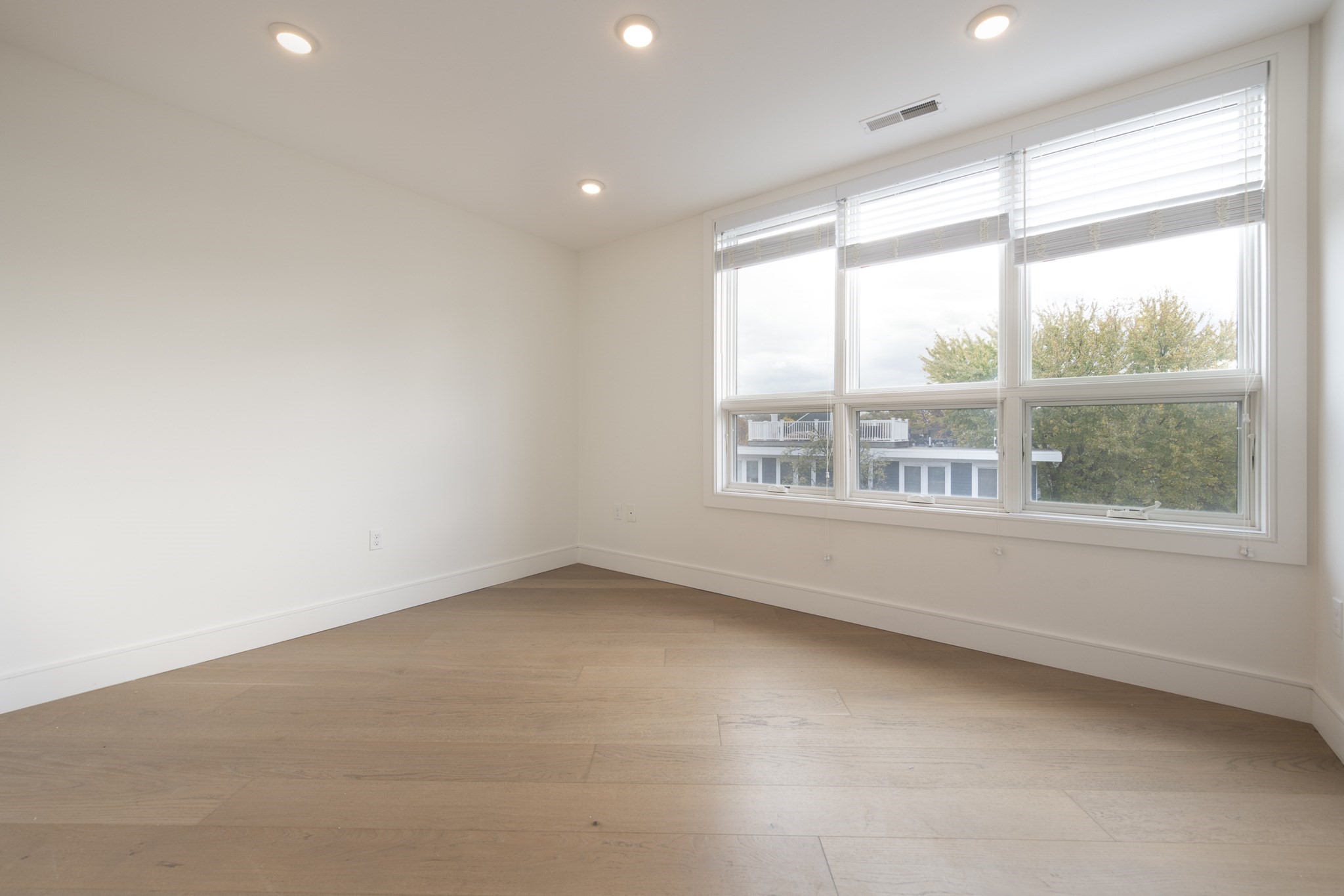 Empty room with light wood floors and a broad bank of windows overlooking autumn trees.
