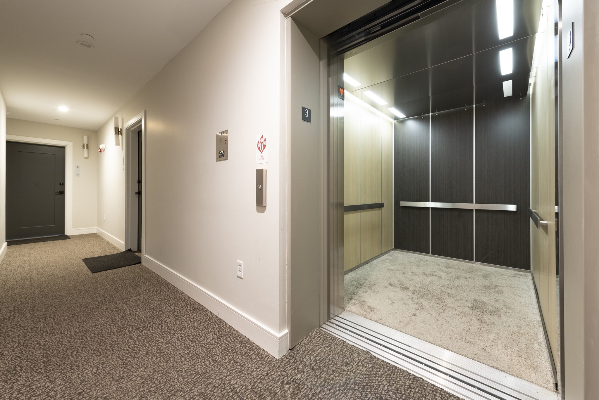 Open elevator with dark wall panels beside a carpeted apartment hallway.