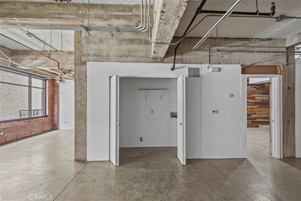 Vacant loft interior with exposed concrete beams, tall industrial windows, brick wall, and open closet.