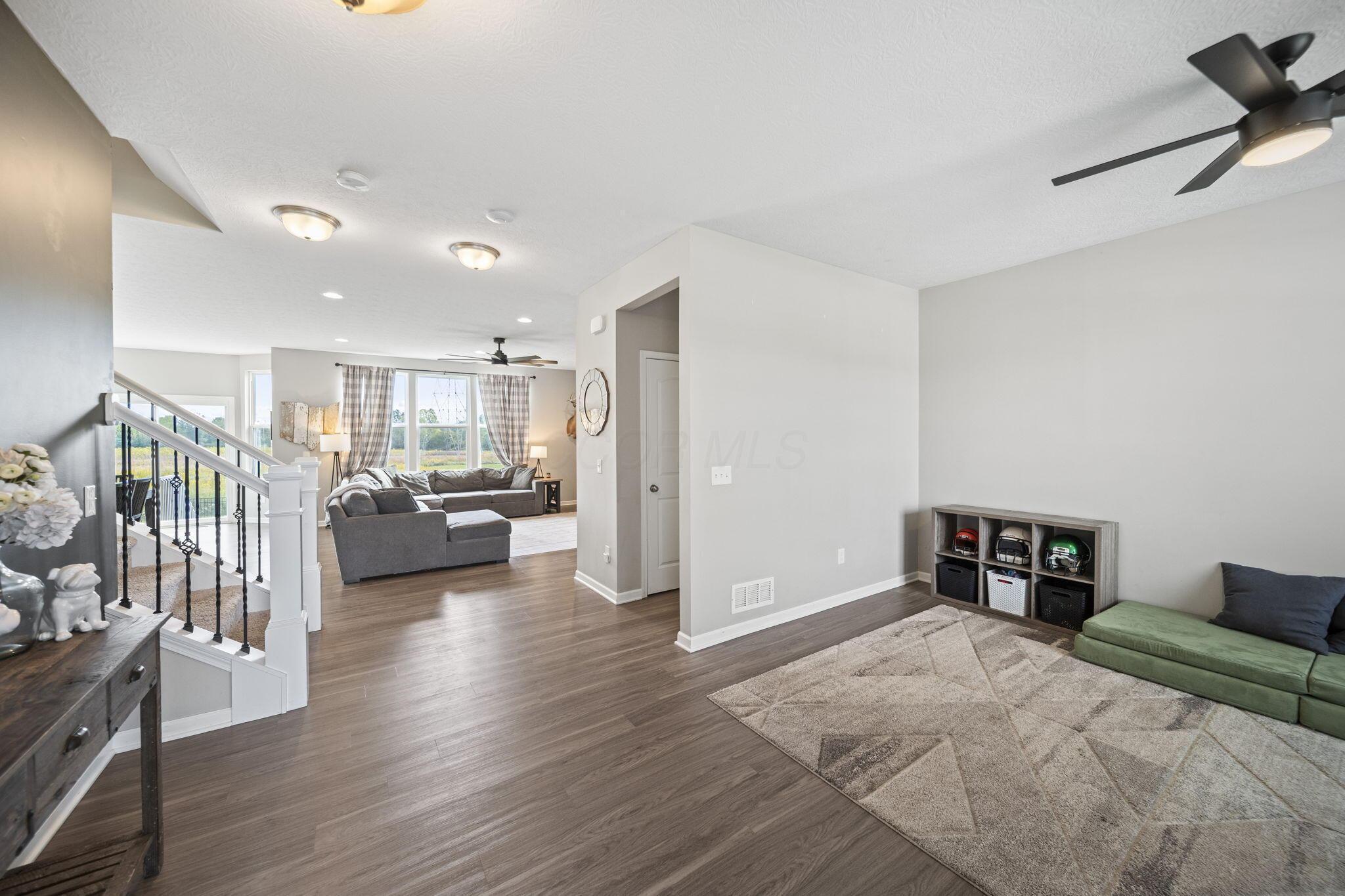 Living room with gray sectional, large curtained windows, open staircase, and green chaise.