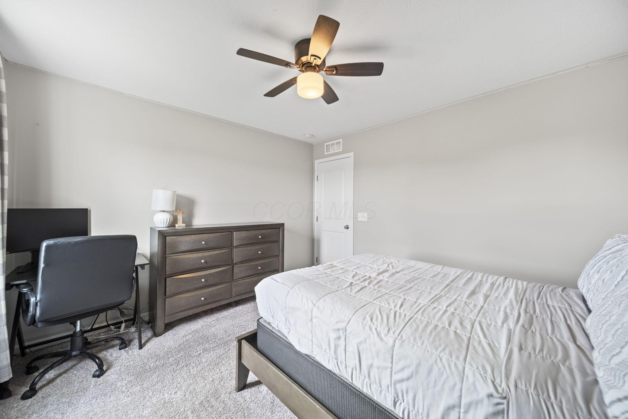 Furnished bedroom with white bedding, a dark dresser, and a desk with computer and office chair.