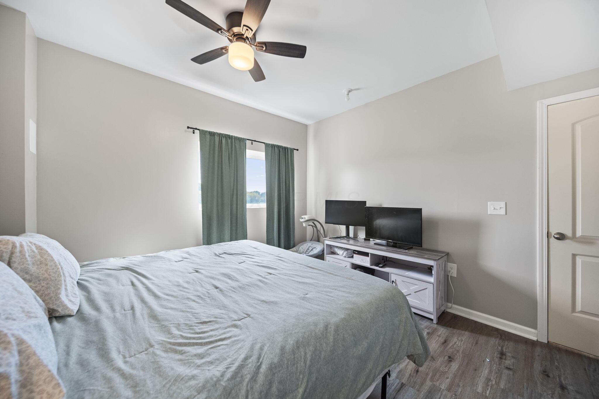 Bedroom with pale gray bedding, green curtains, and two monitors on a console.