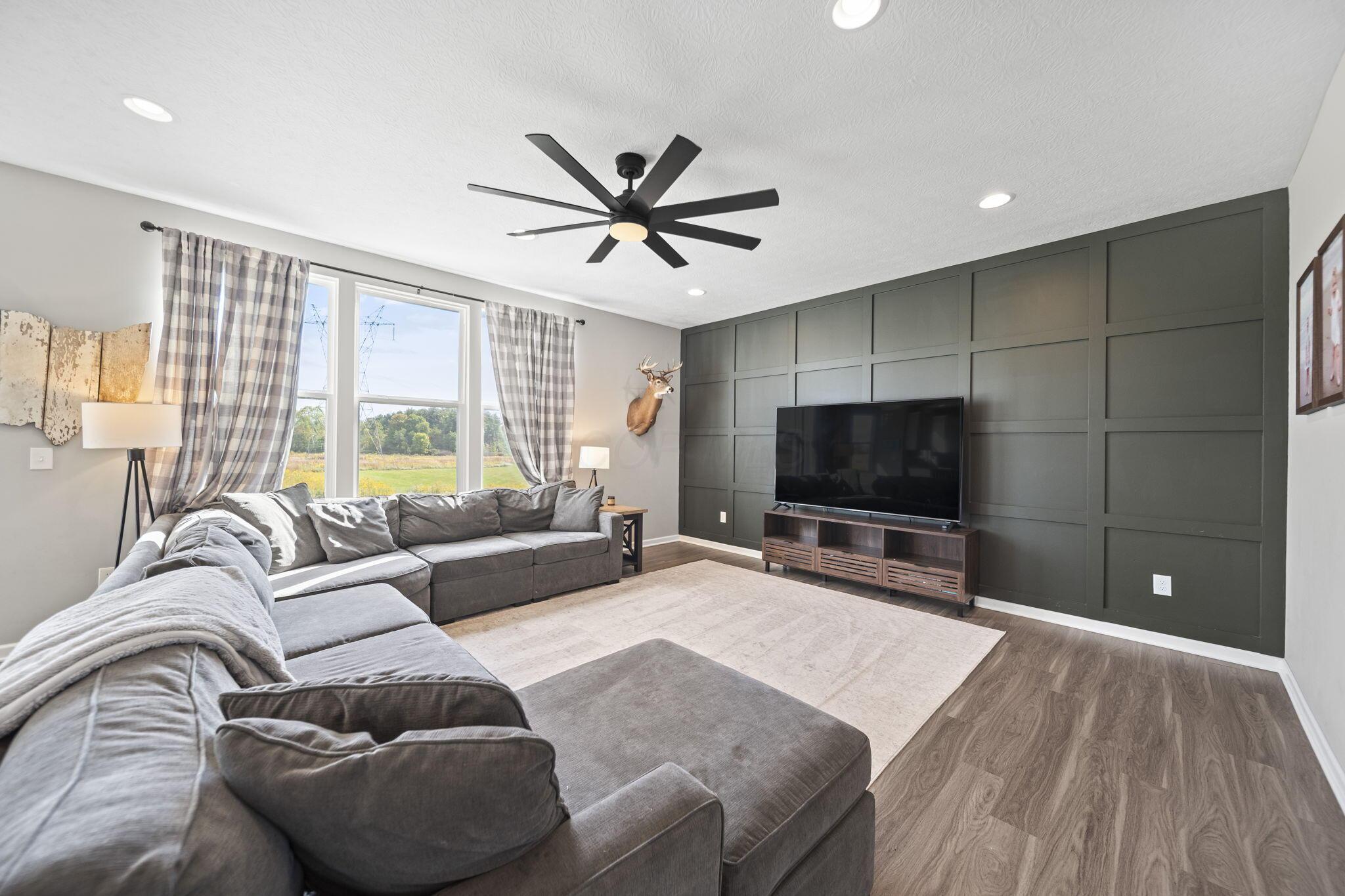 Living room with a gray sectional, dark paneled TV wall, and windows overlooking a grassy yard.