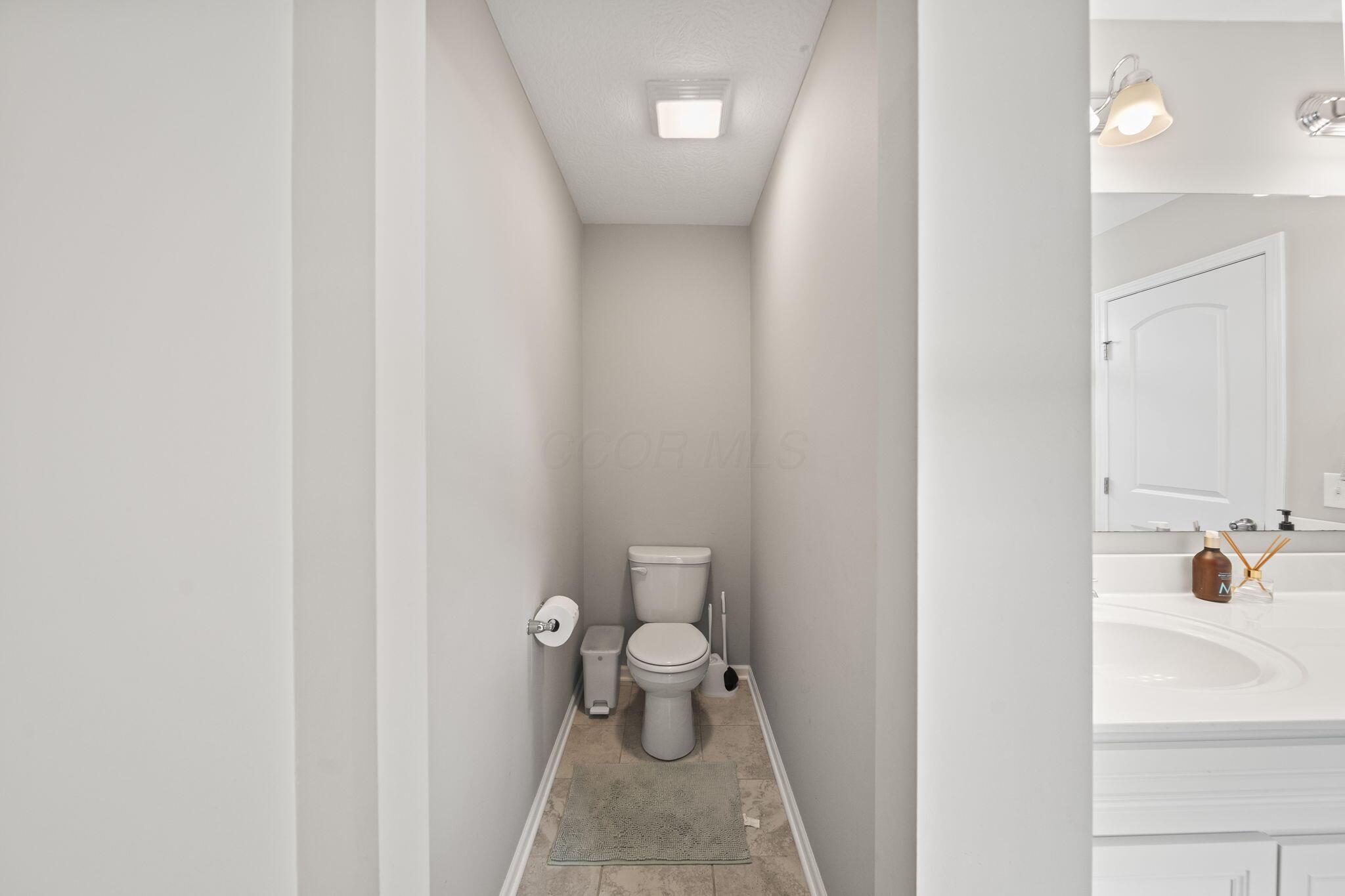 Narrow toilet alcove with a white toilet, tiled floor, and part of a bathtub and sink.