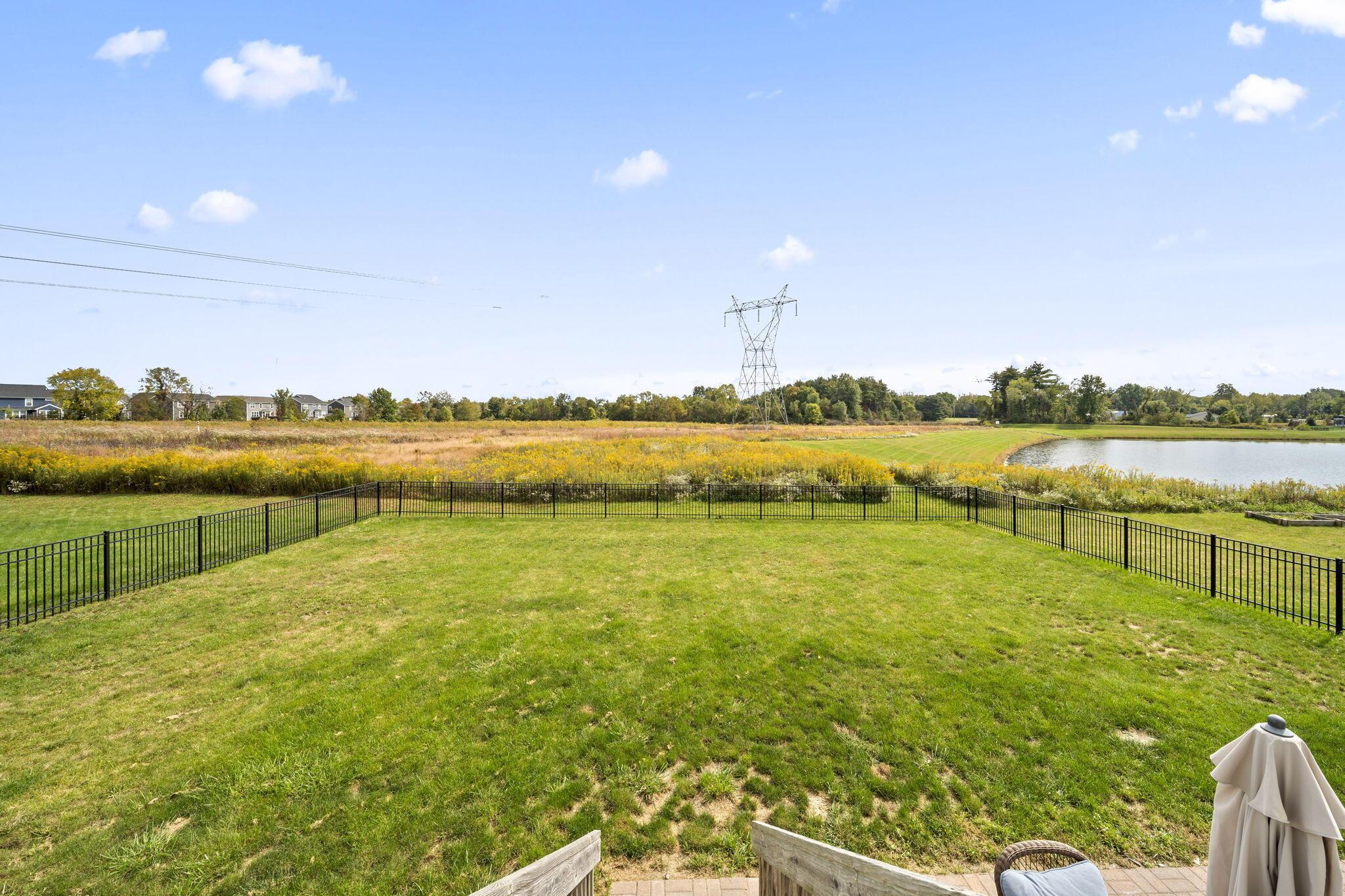 Fenced backyard lawn overlooking a pond and yellow wildflower field, with a transmission tower in the distance.