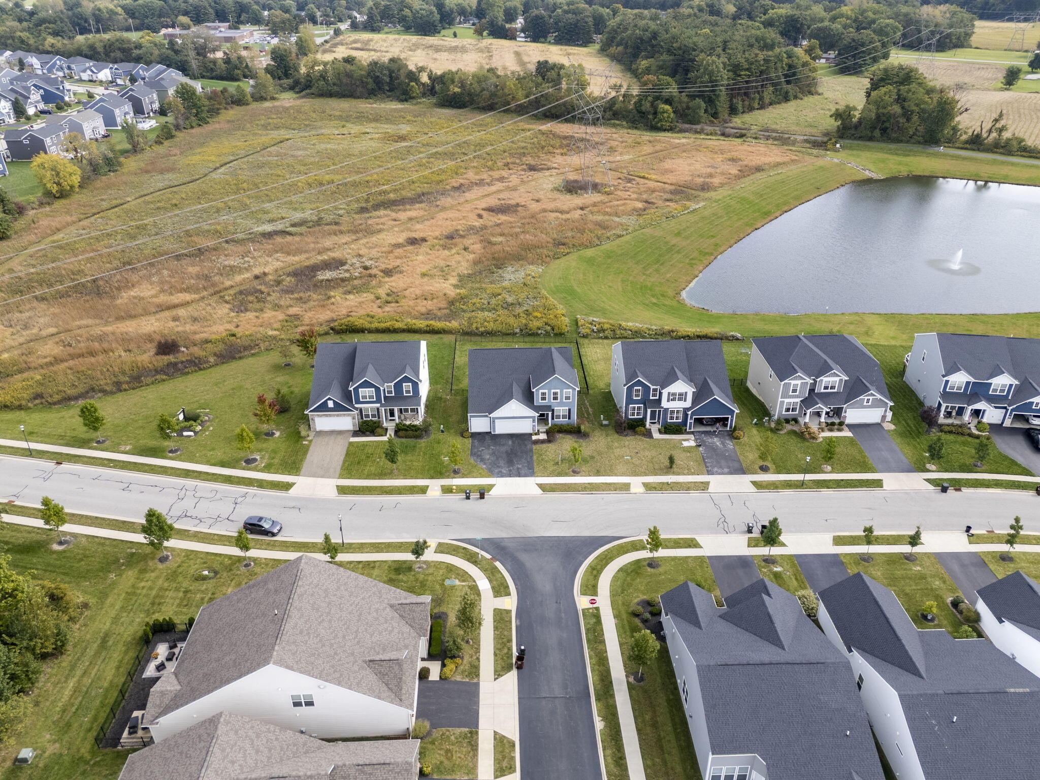 Aerial neighborhood view with detached homes, a pond fountain, and open fields crossed by power lines.