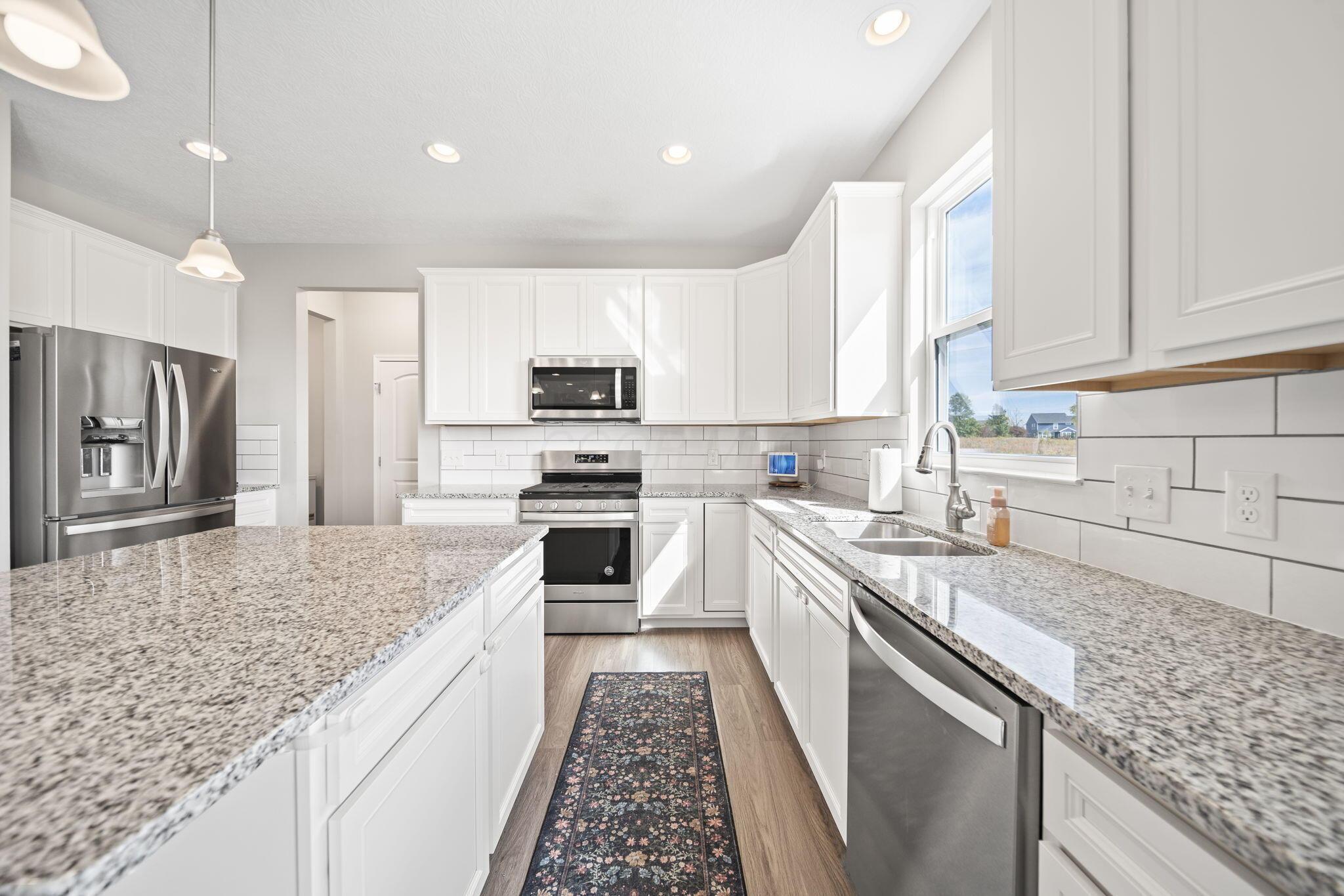 Bright white kitchen with granite island, stainless steel appliances, and a sink beneath a sunny window.