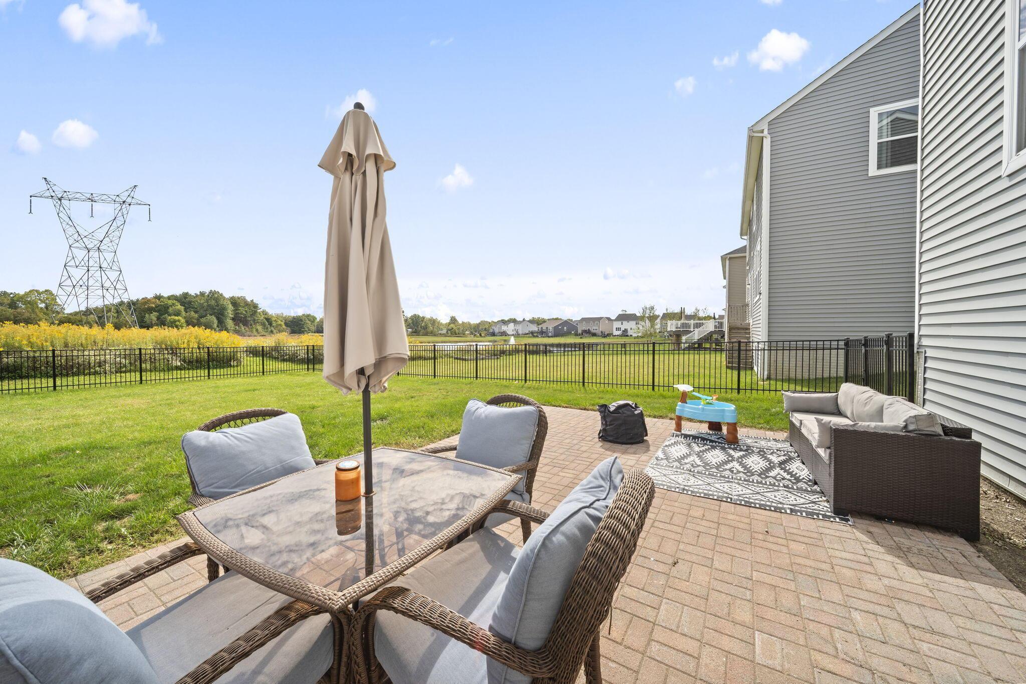 Brick patio with wicker seating and a closed umbrella, overlooking a fenced lawn and open field.