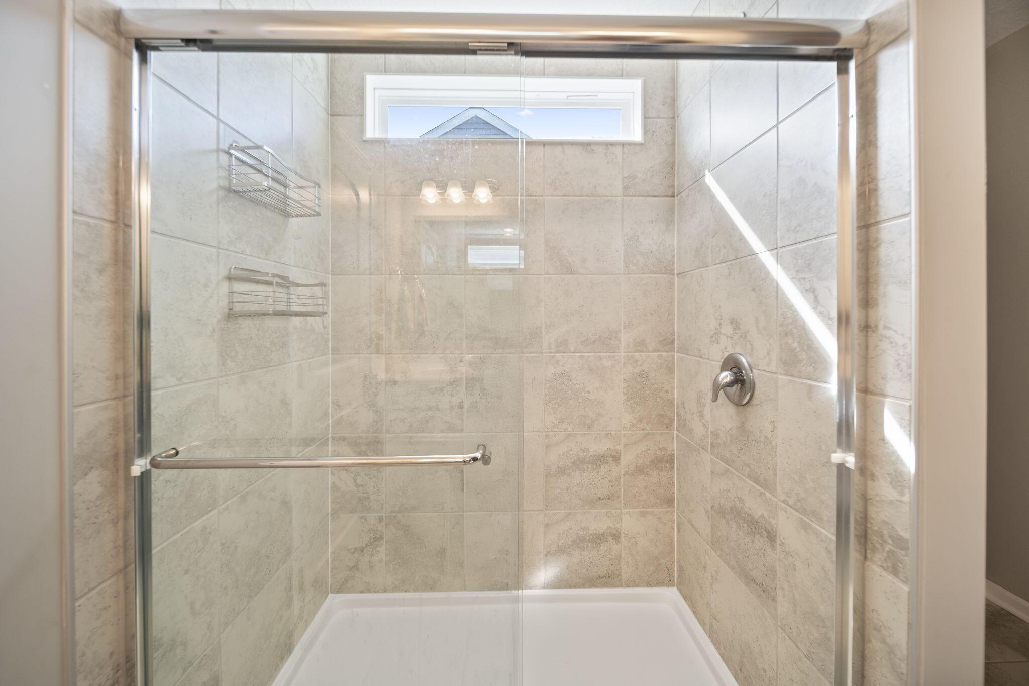 Bathroom shower with beige tile, glass doors, chrome fixtures, and a high narrow window.
