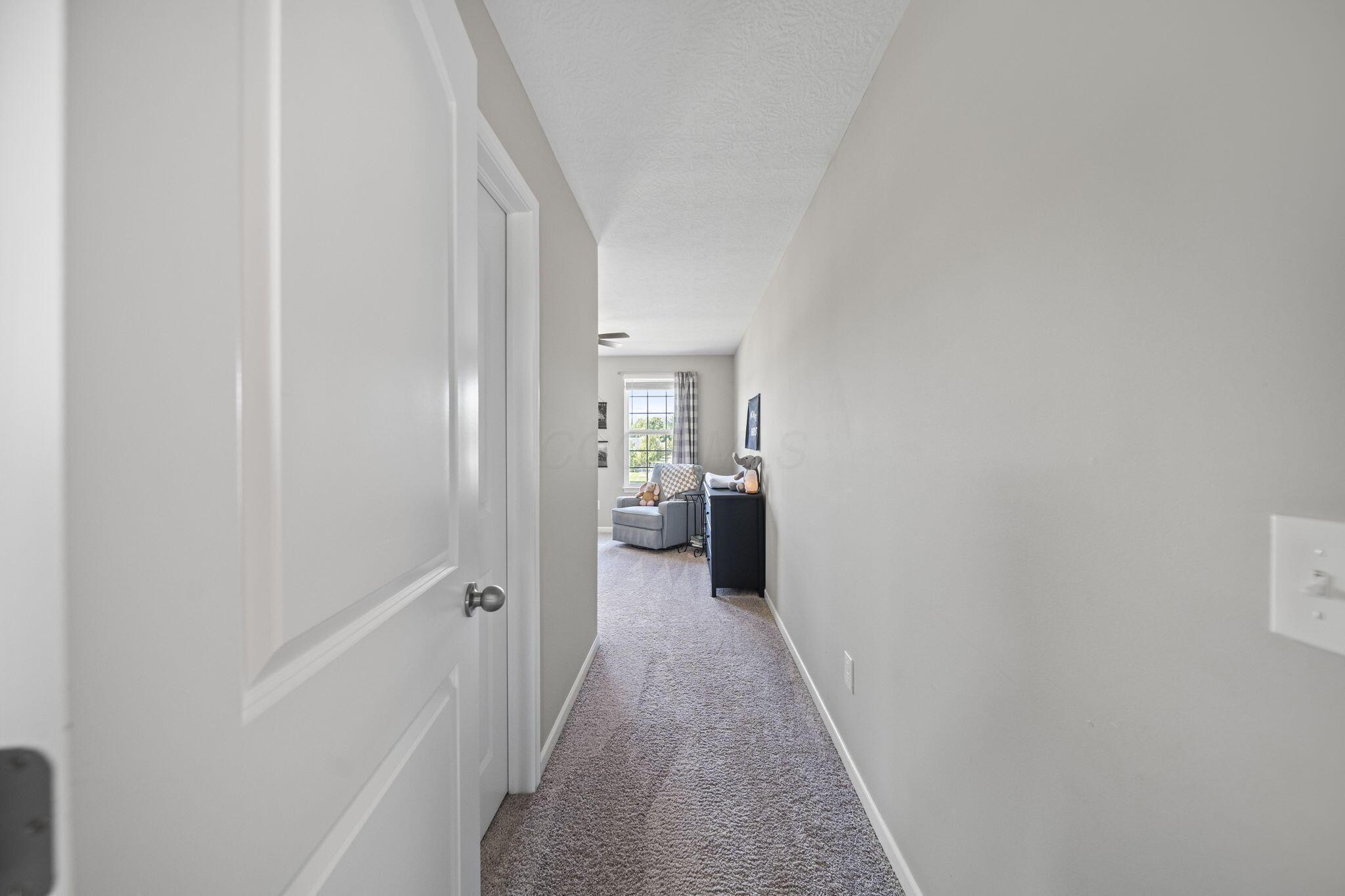 Carpeted hallway opening onto a furnished room with a gray armchair, stuffed toys, and a window.