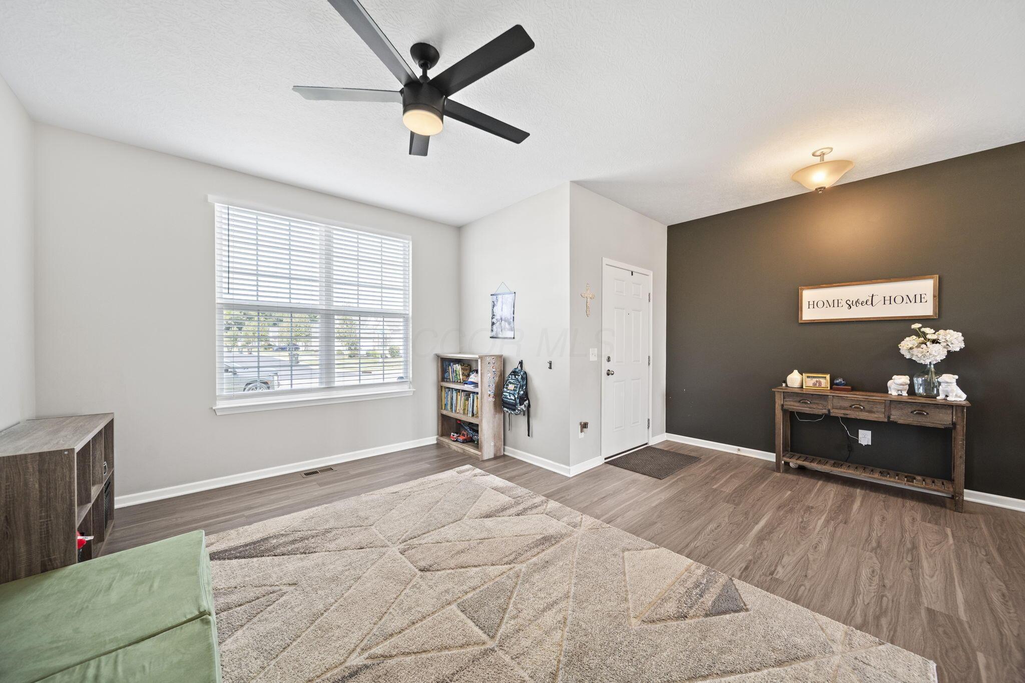 Living room with a dark accent wall, geometric rug, ceiling fan, and large window with blinds.
