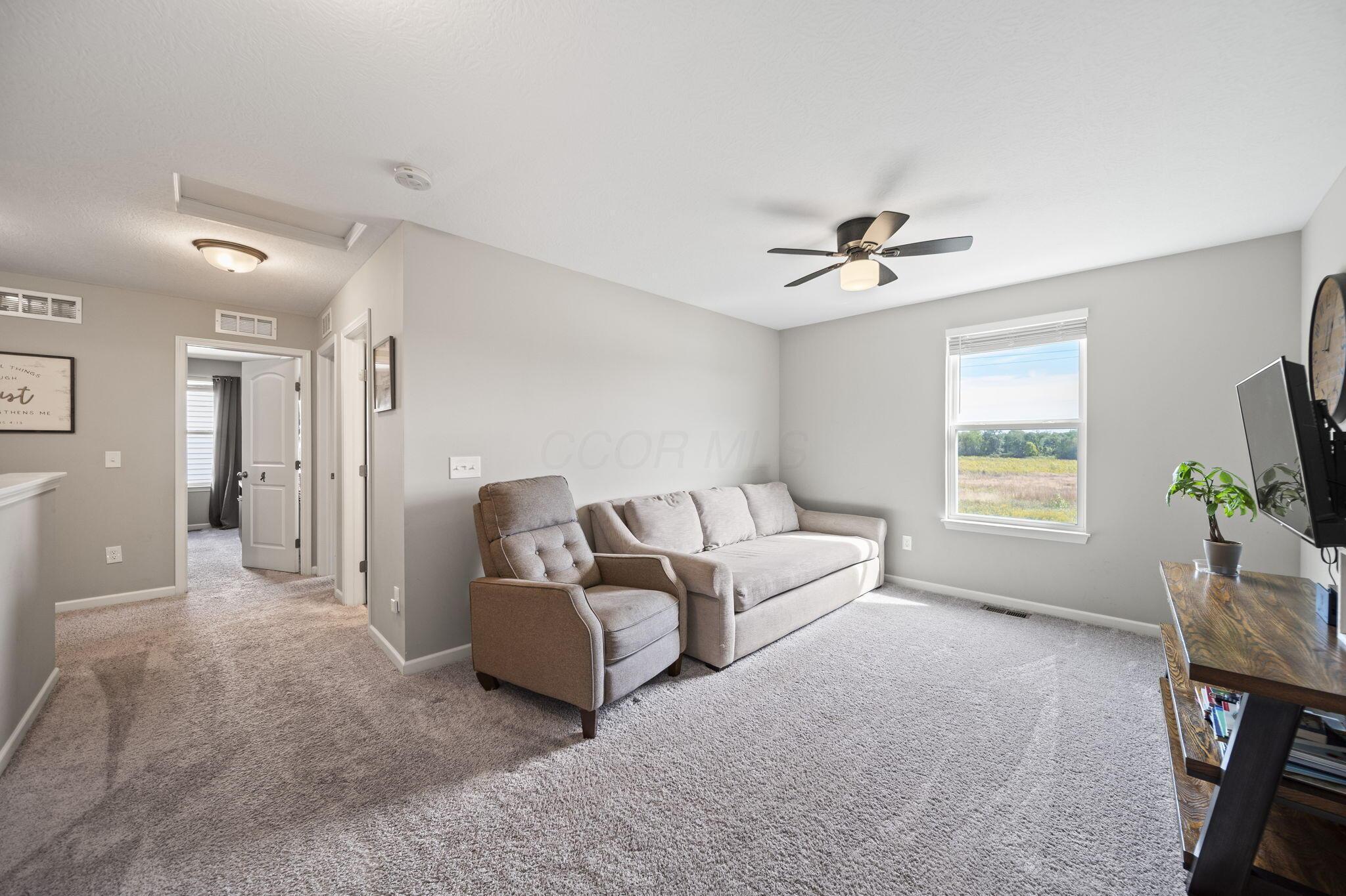Furnished living room with a beige sofa and armchair, ceiling fan, and window overlooking a grassy field.